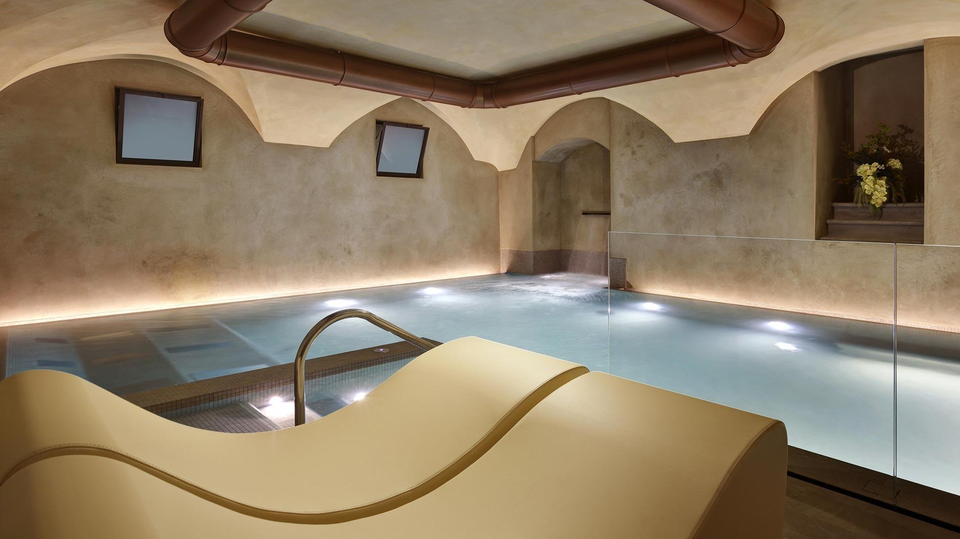 an underground spa pool lit from below, with a vaulted stone ceiling, exposed copper pipework, and a cream leather lounger in the foreground