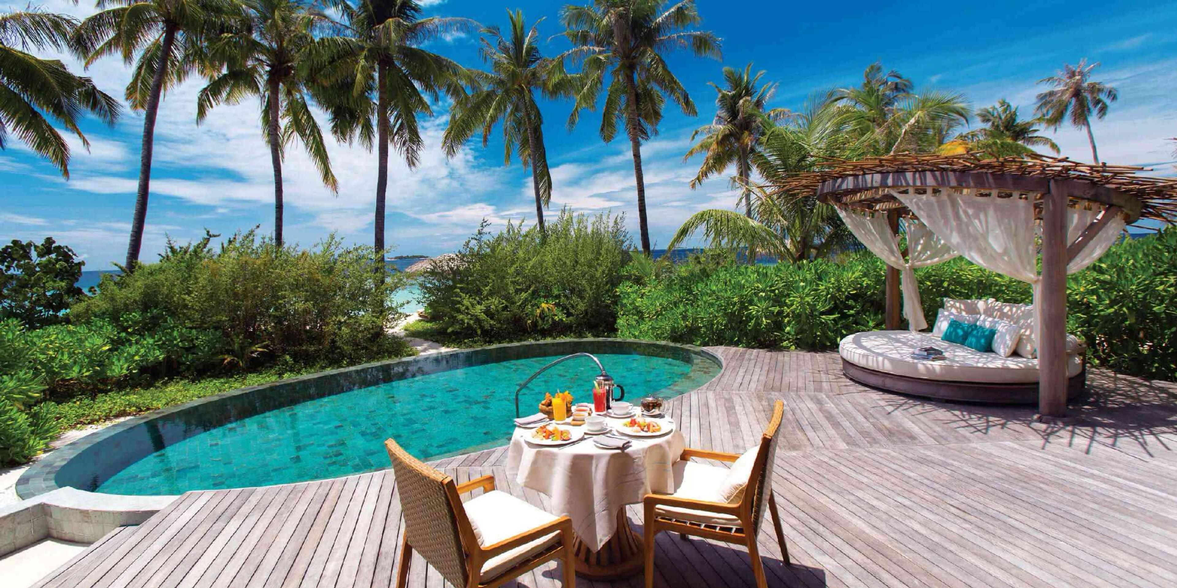 A breakfast table set with plates and juice beside a curved pool, with a shaded daybed and palm trees around the deck.