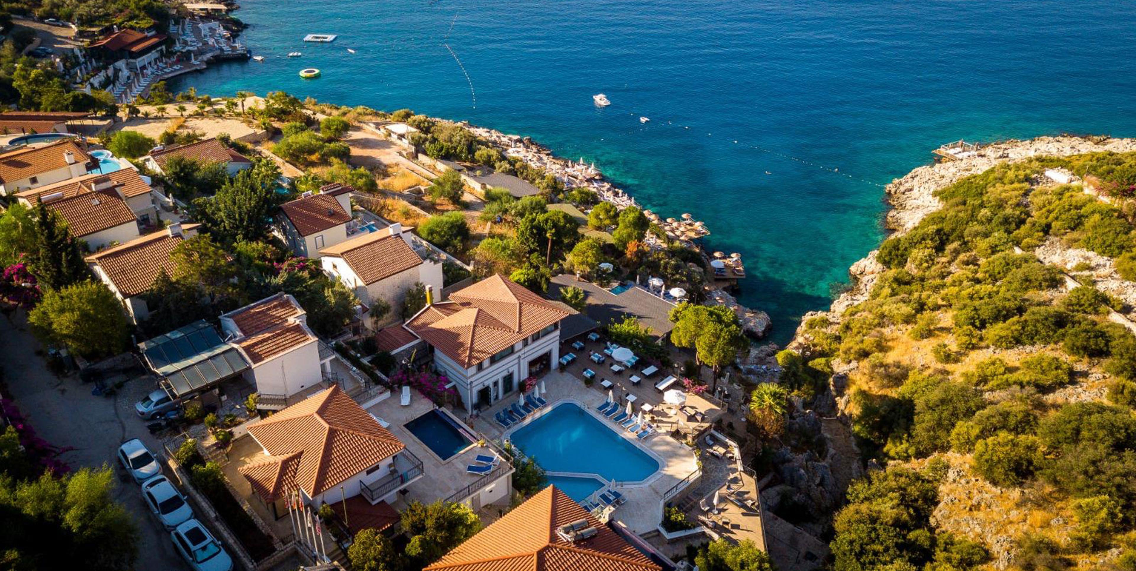 An aerial view of a hotel with terracotta roofs and a swimming pool set on a cliff above a rocky turquoise bay, with boats moored offshore.
