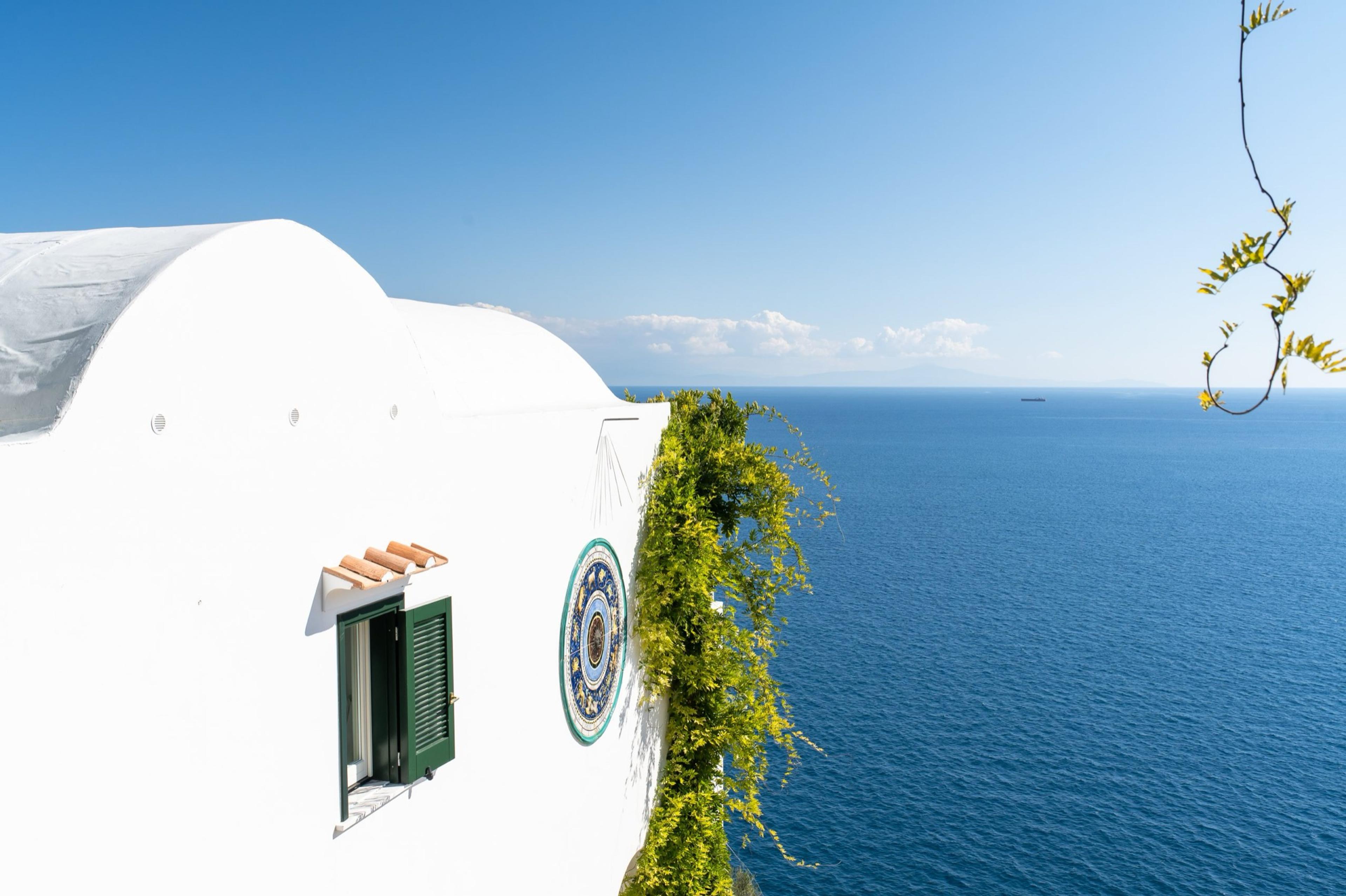 The white rendered corner of a cliffside building with a ceramic plate set into the wall and a vine trailing down against the sea.