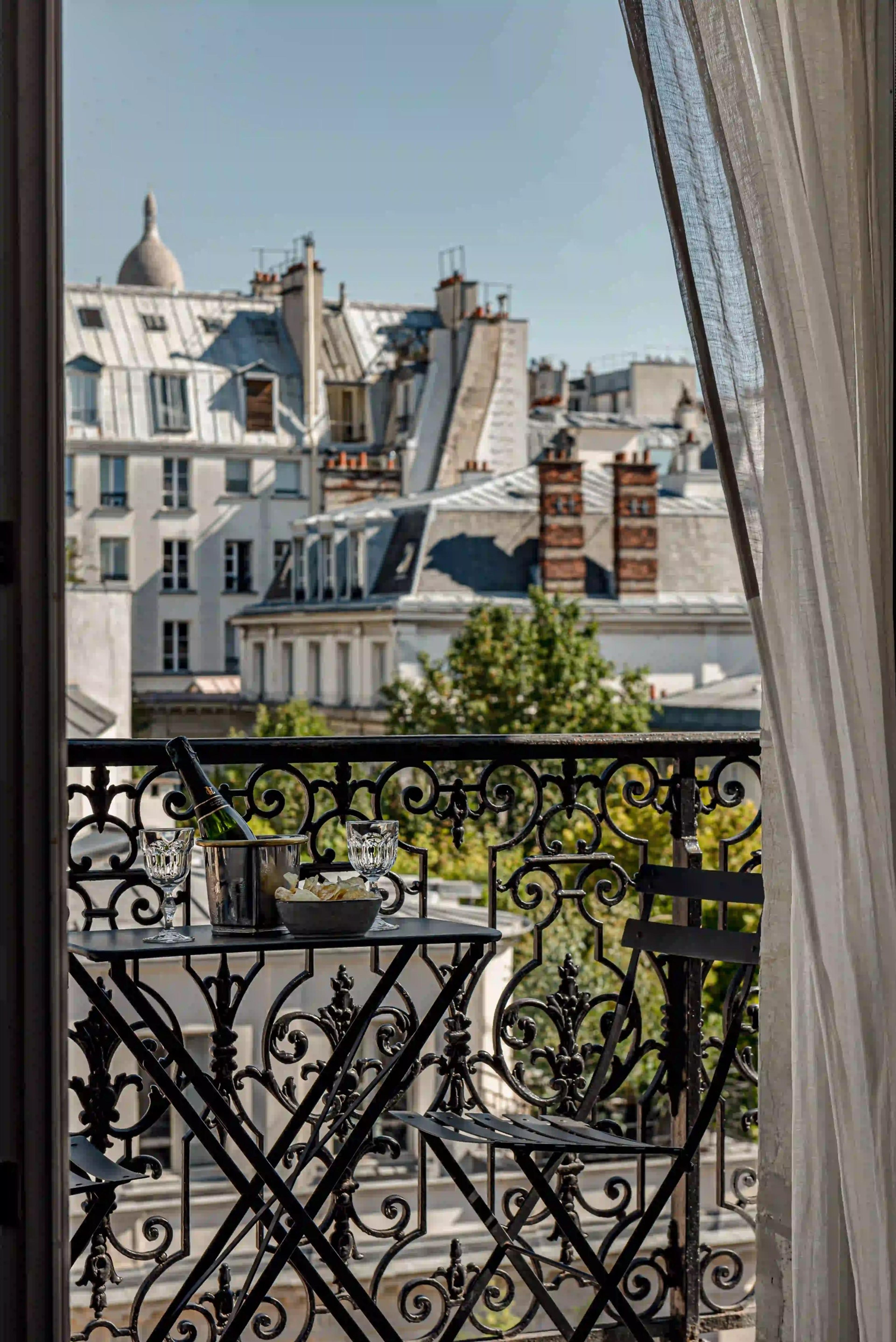 A small balcony table set with a champagne bottle, two glasses and a bowl of snacks looks out over Paris rooftops and the dome of Sacre-Coeur.