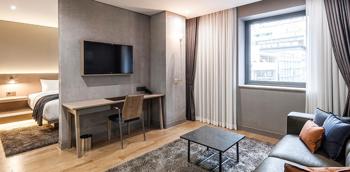 A corner hotel suite with a bed on a raised platform to one side, a desk and wall-mounted television, and a sofa facing a large window onto the city.