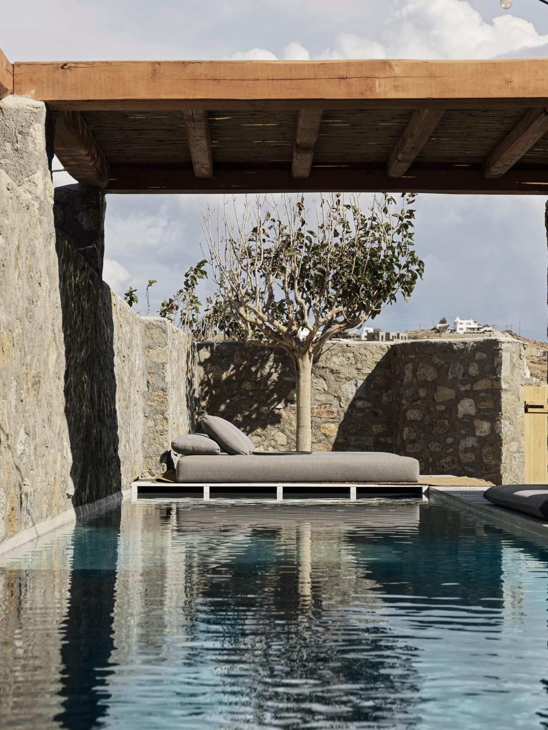 A single grey daybed sits on a stone pool deck beneath a wooden pergola, a bare tree and stone walls behind it reflected in the still water.