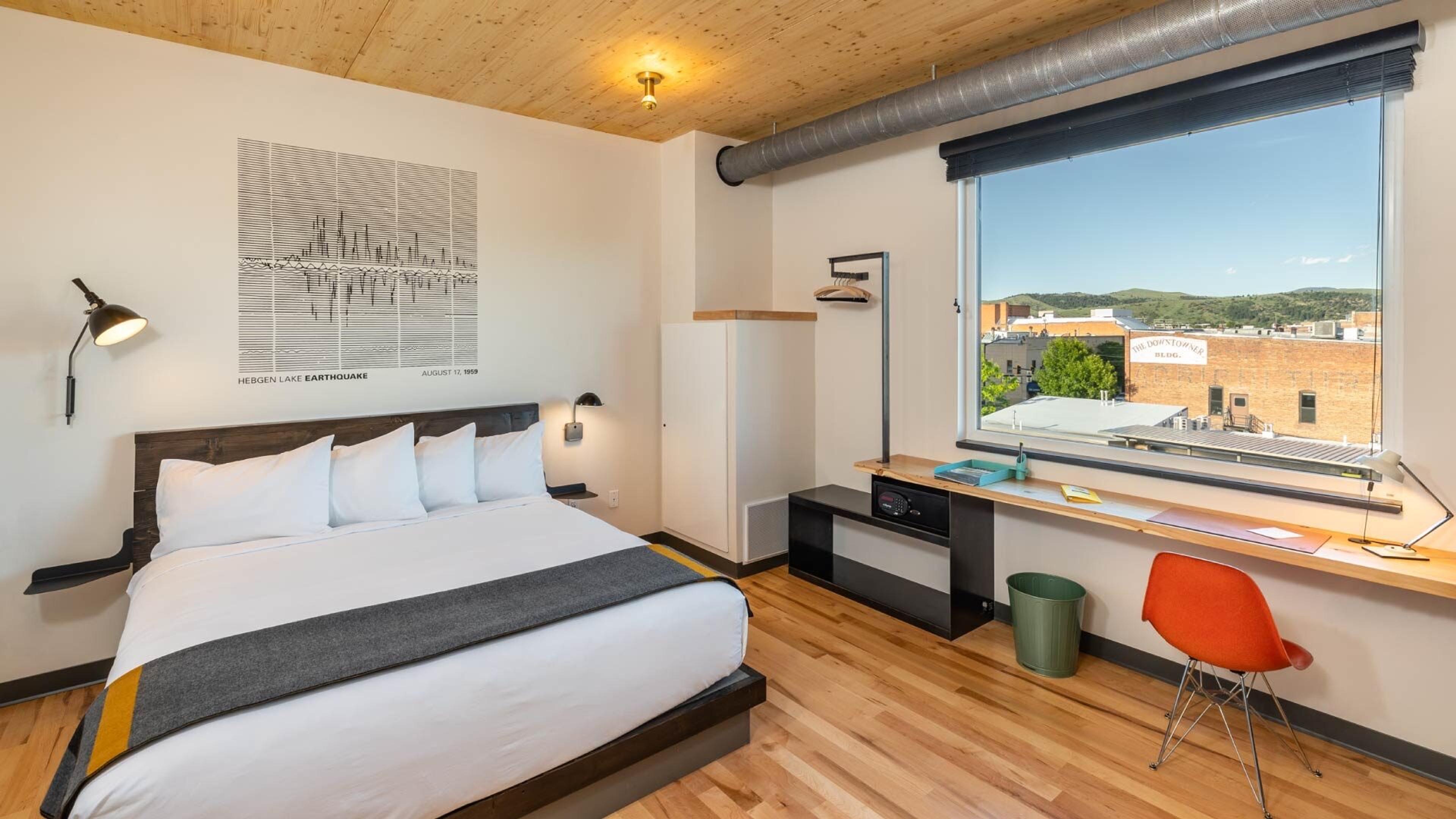 A king bed with white linen and a grey and yellow throw, a desk under a window looking over rooftops and hills, and an orange chair.