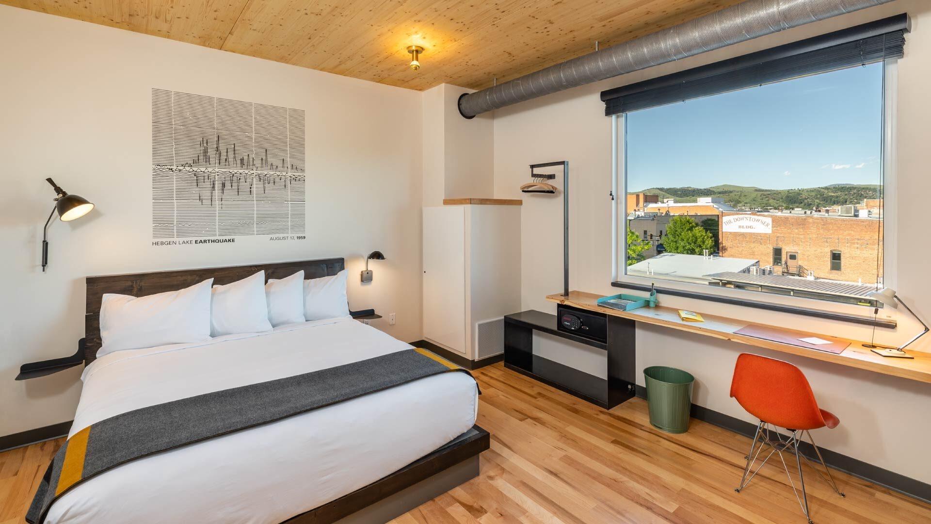 A king bed with white linen and a grey and yellow throw, a desk under a window looking over rooftops and hills, and an orange chair.