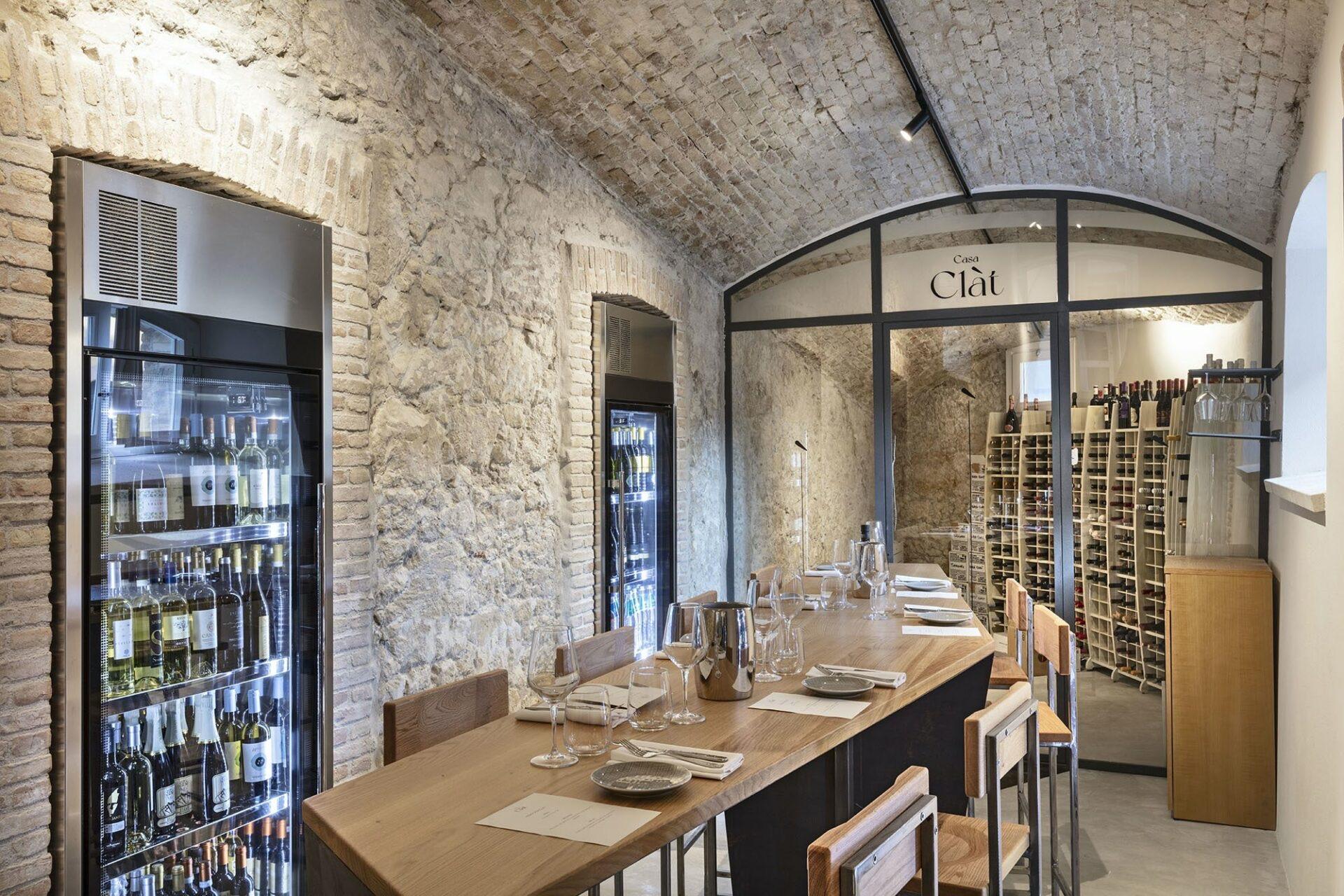 A vaulted brick wine cellar with a long wooden table set for a meal, wine fridges built into the stone wall, and floor-to-ceiling wine racks beyond a glass door marked Casa Clat.