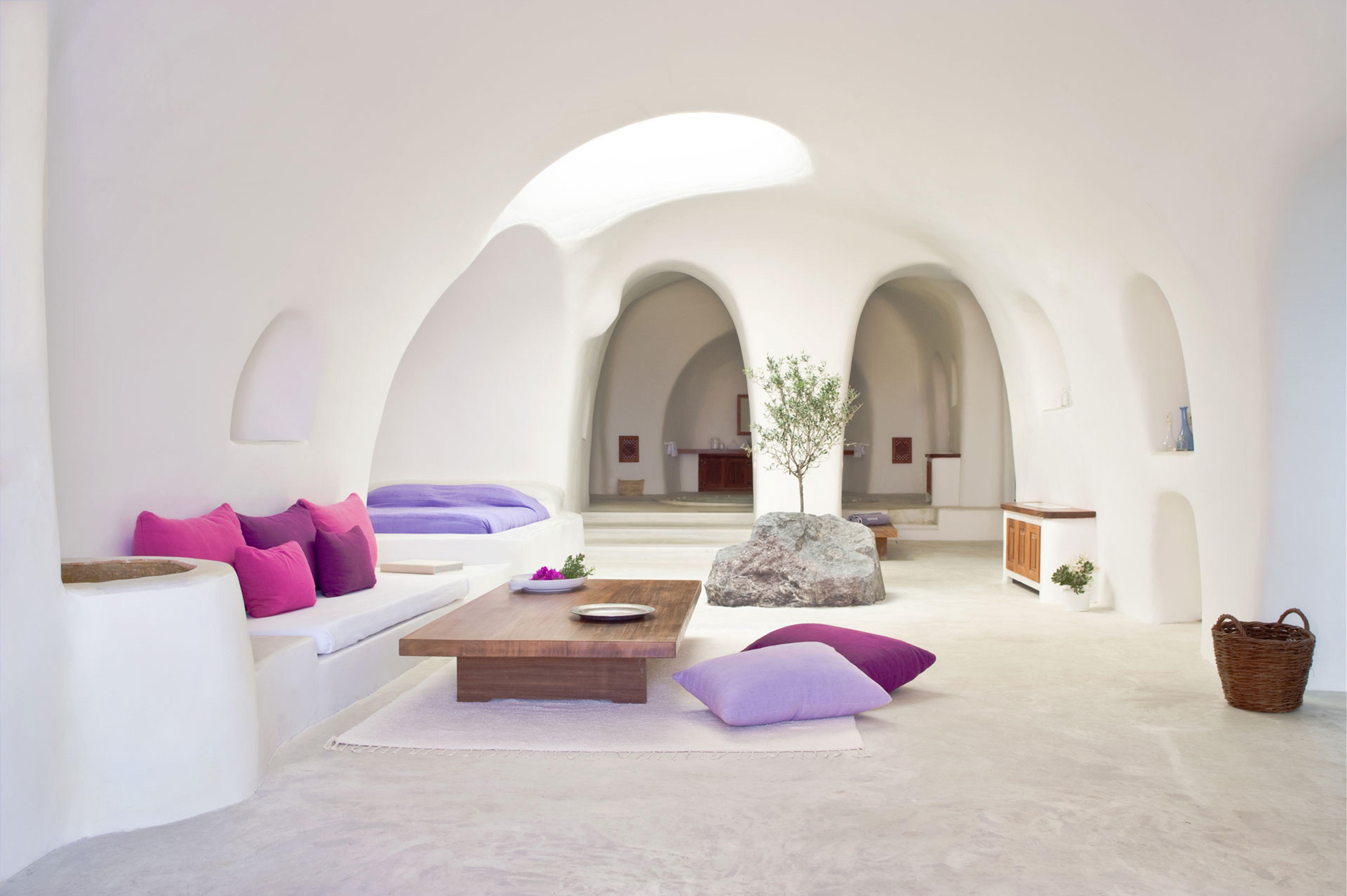 A curved whitewashed cave suite with pink cushioned seating, a low wooden table, a boulder beside a small olive tree, and archways leading to a bathroom.