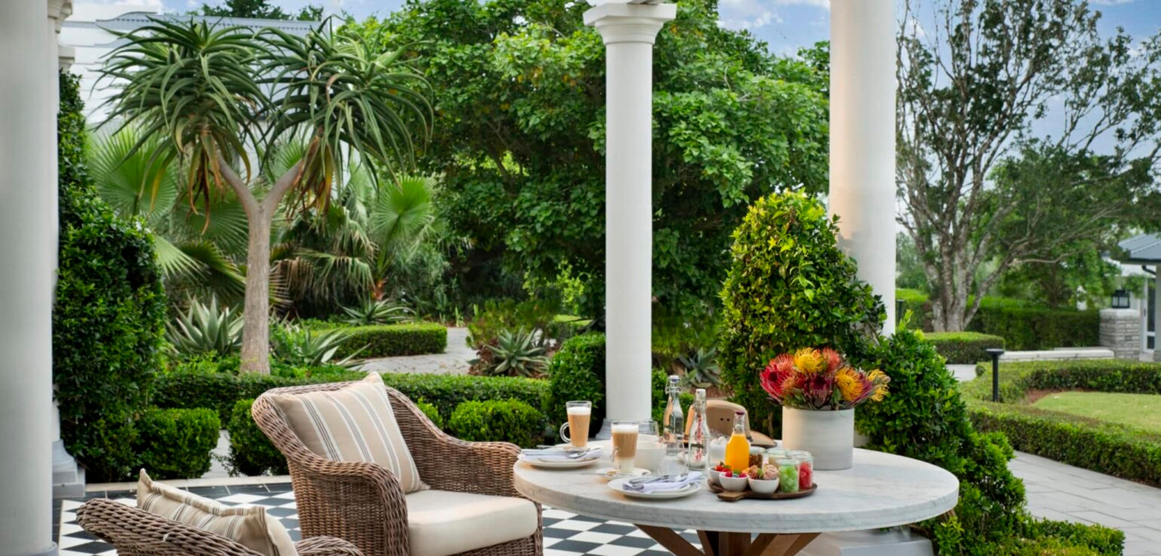A round table laid for breakfast with coffee, juice and fruit on a black and white checkered verandah, between white columns.