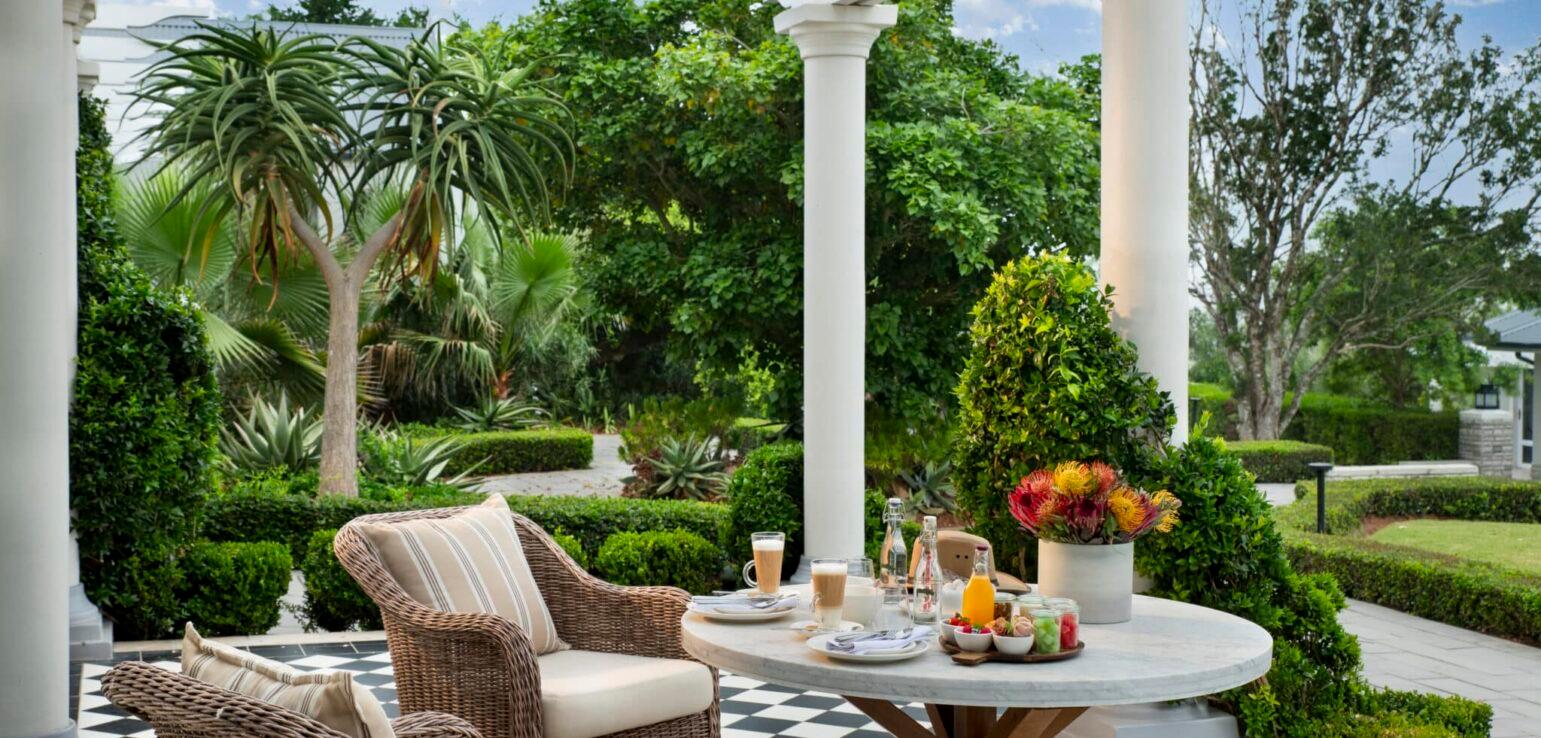A round table laid for breakfast with coffee, juice and fruit on a black and white checkered verandah, between white columns.