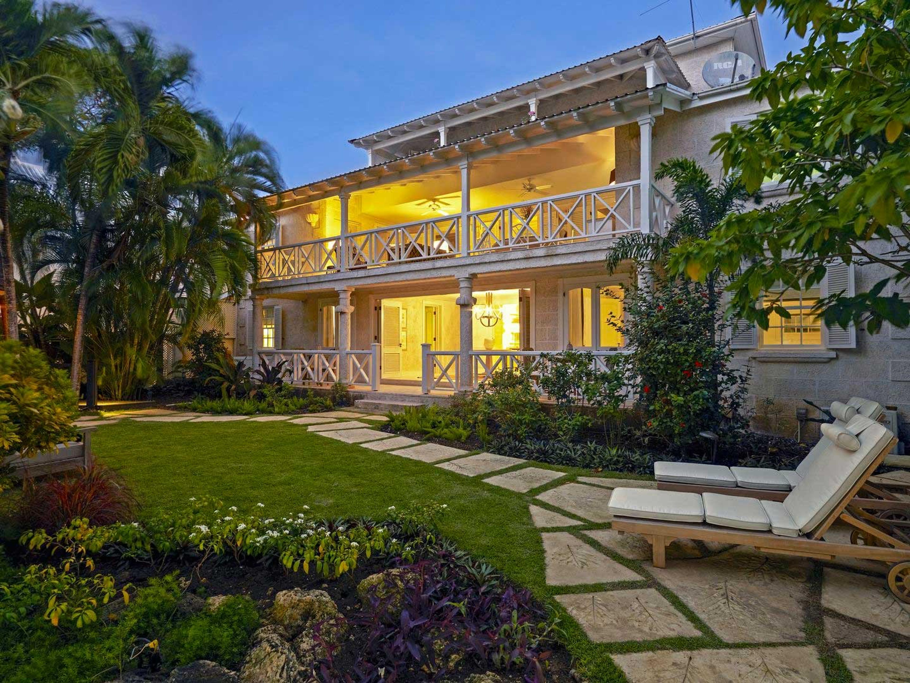 A two-storey house lit from within at dusk, seen across a lawn and flowerbeds with a sun lounger in the foreground