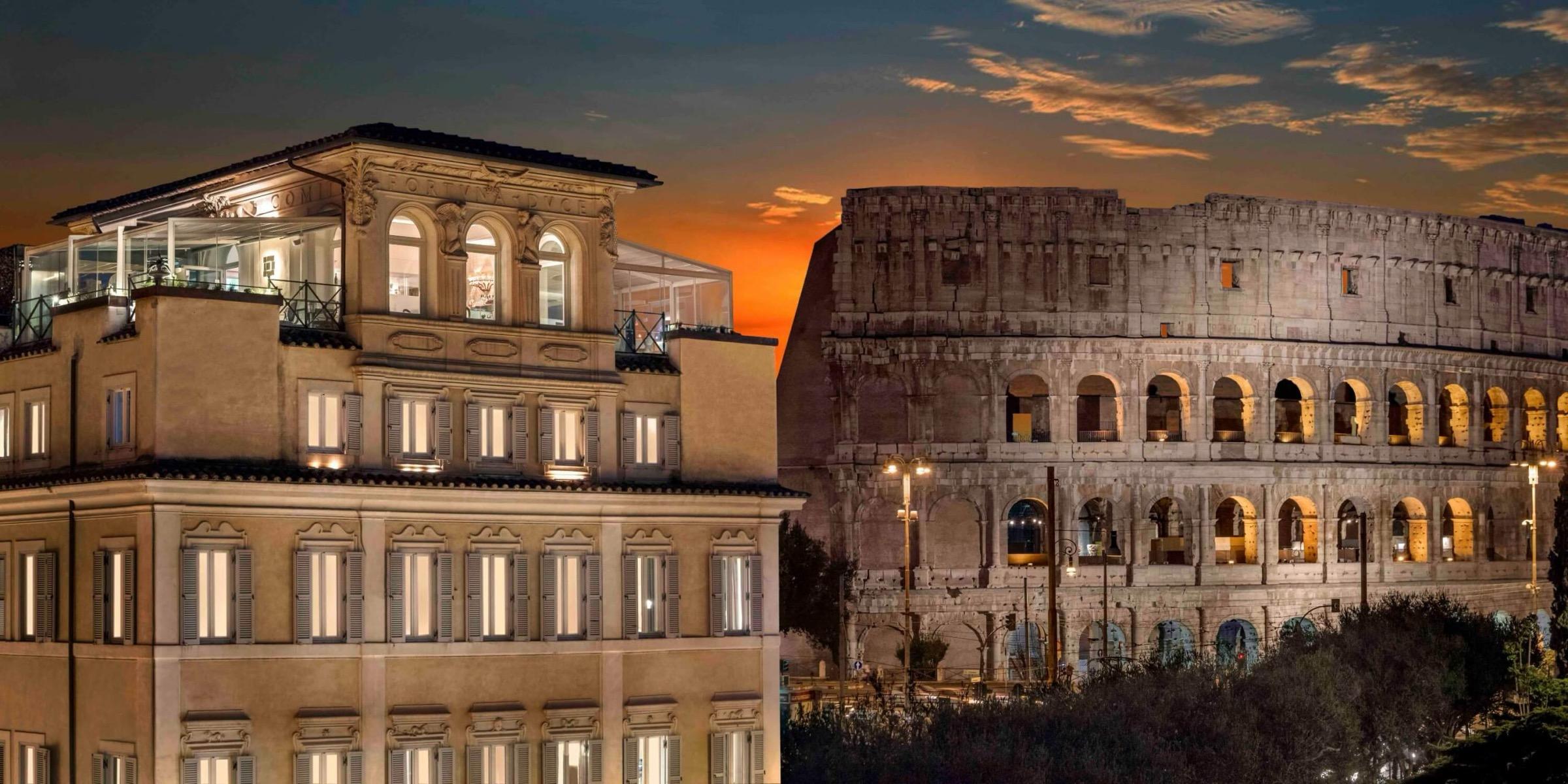 A cream-coloured palazzo at dusk, its lit windows and top-floor glass terrace in front, with the Colosseum rising behind it to the right.