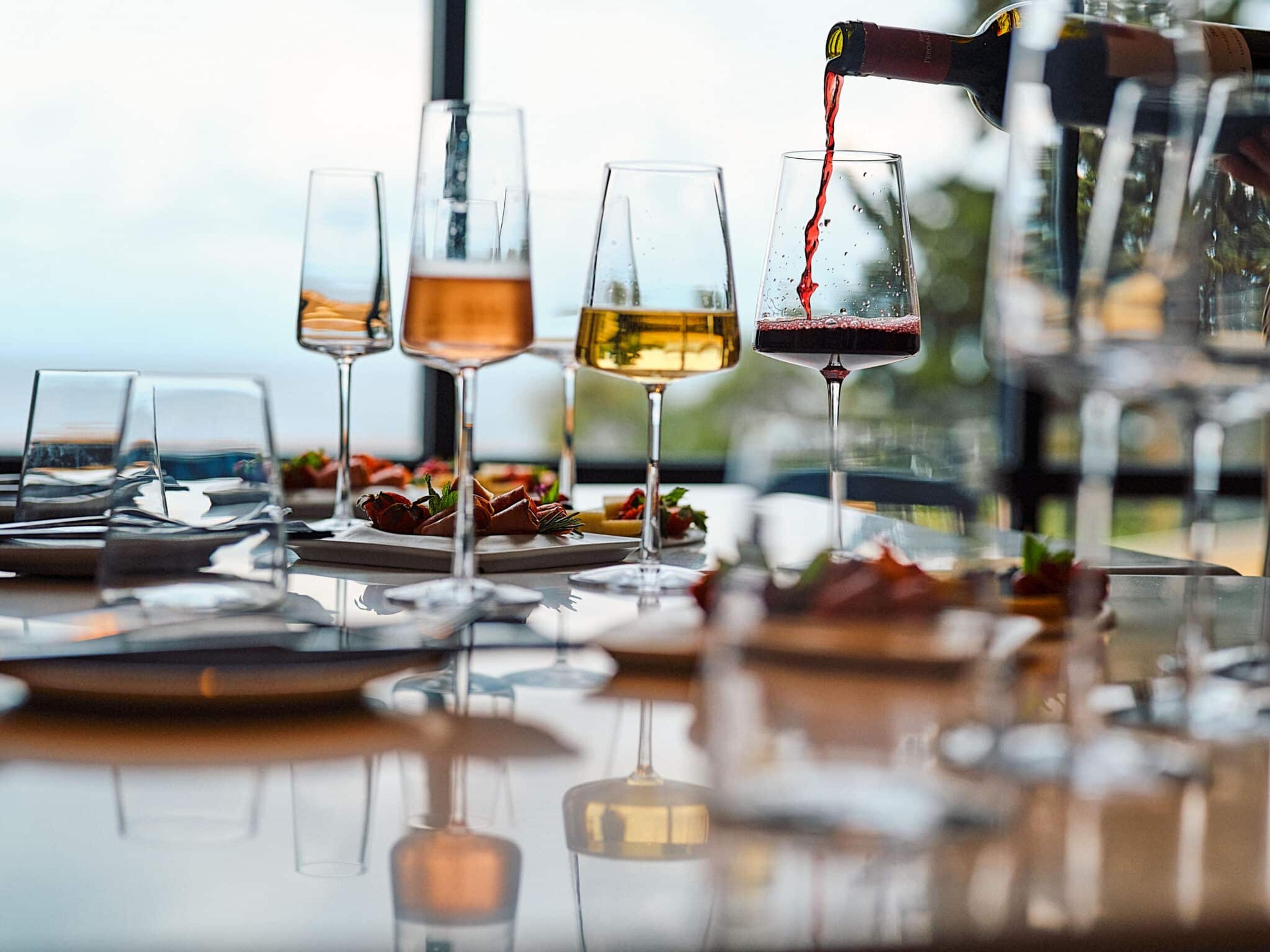 Red wine is poured into a glass on a table set with white wine, rosé and small plates of food, a blurred sea view behind.