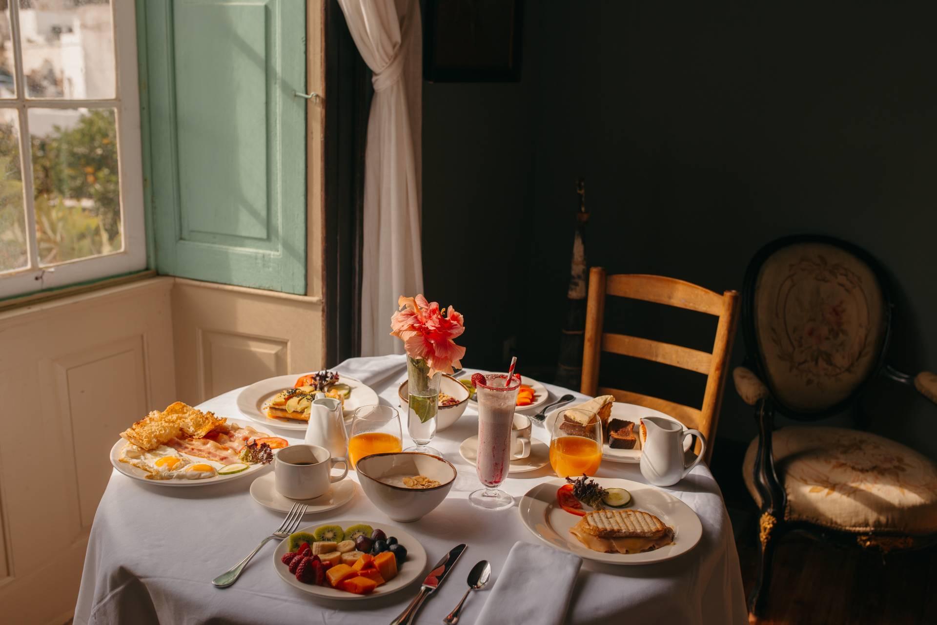 A breakfast spread of pastries, fruit, coffee and juice laid on a round table beside an open window with green shutters.