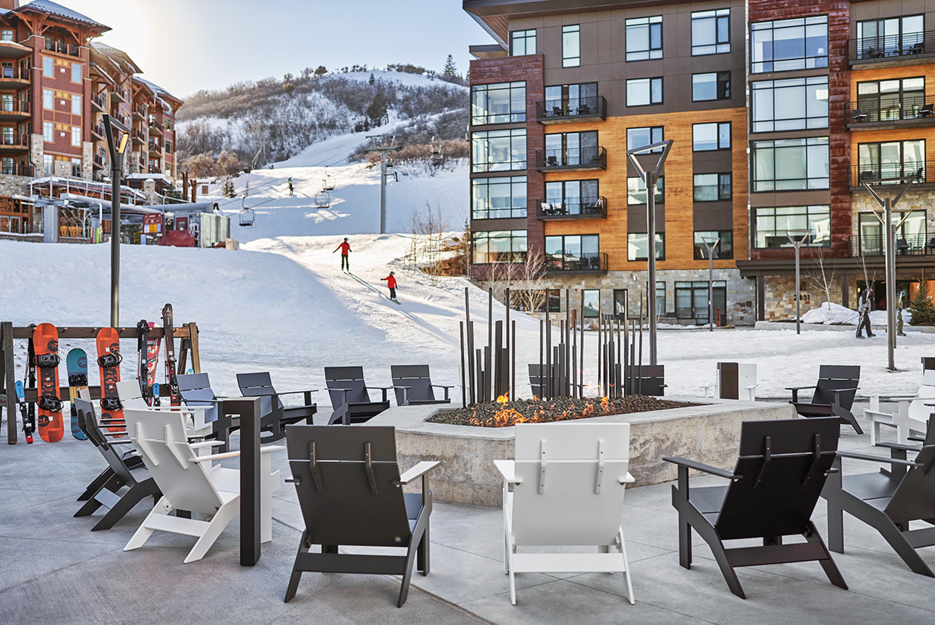 A paved terrace with grey Adirondack chairs around a round concrete fire pit, a rack of snowboards at the left, a slope with two skiers in the distance, and a multi-storey hotel building beside it.