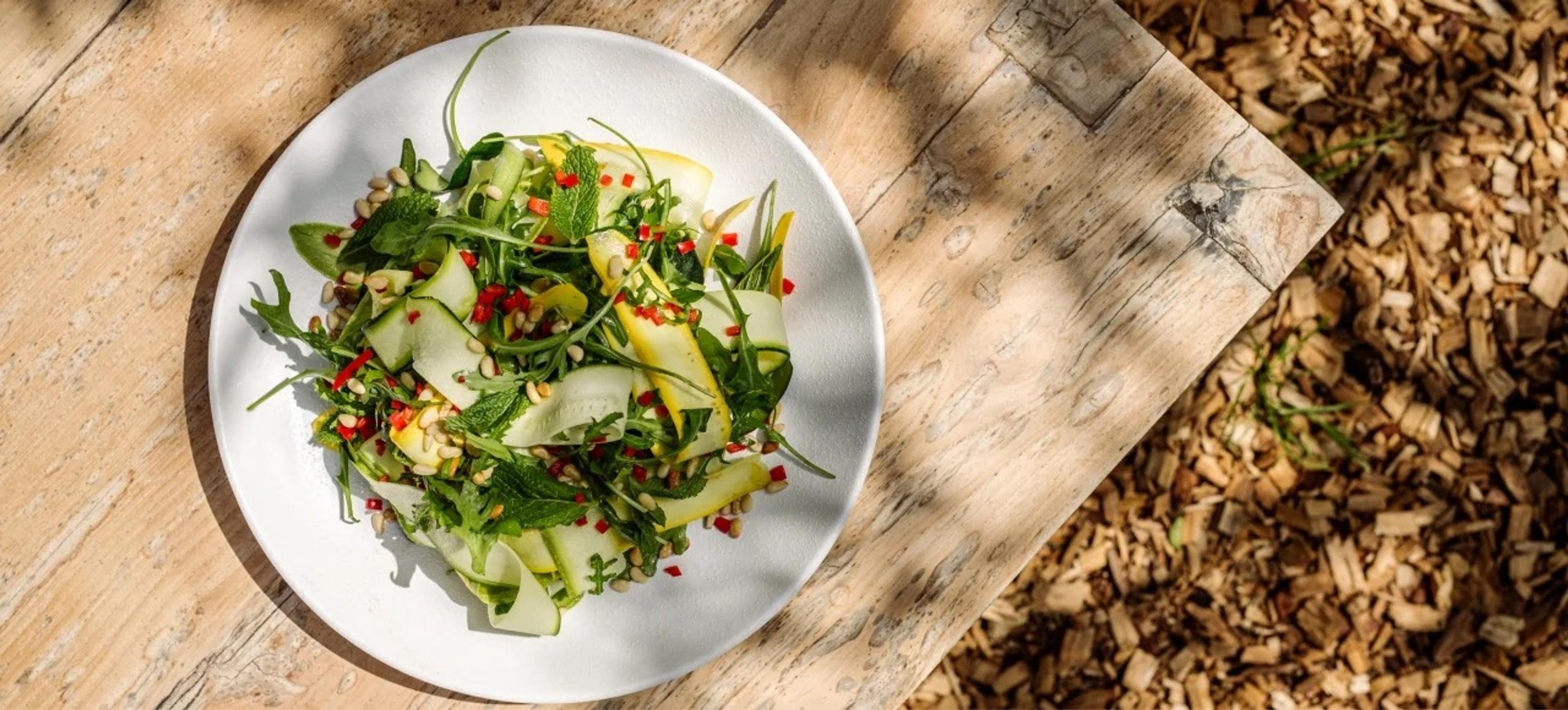 An overhead view of a plate of shaved courgette and mango salad with rocket, mint, chilli and seeds, on a rustic wooden table.