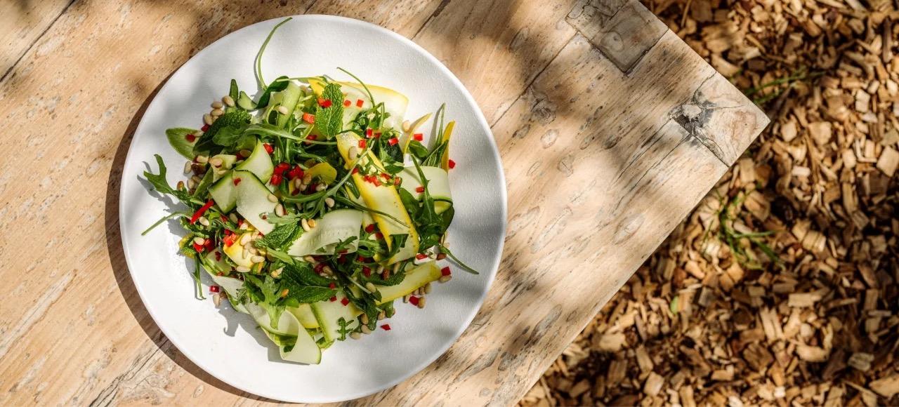 An overhead view of a plate of shaved courgette and mango salad with rocket, mint, chilli and seeds, on a rustic wooden table.