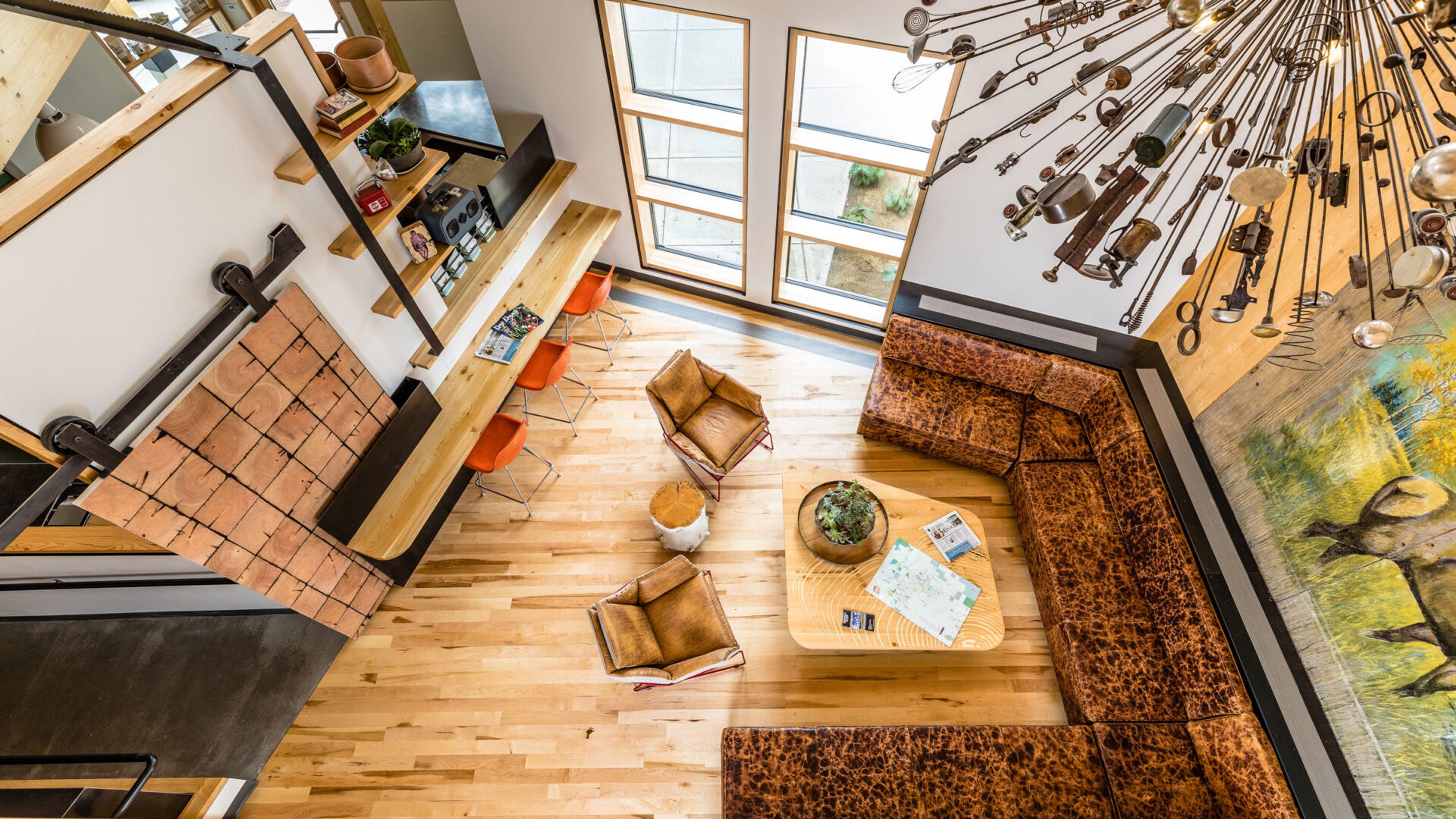 Looking down from an upper level onto a lobby with a leather sectional, a low wooden table, a tiled fireplace on the left and a chandelier made of metal tools.