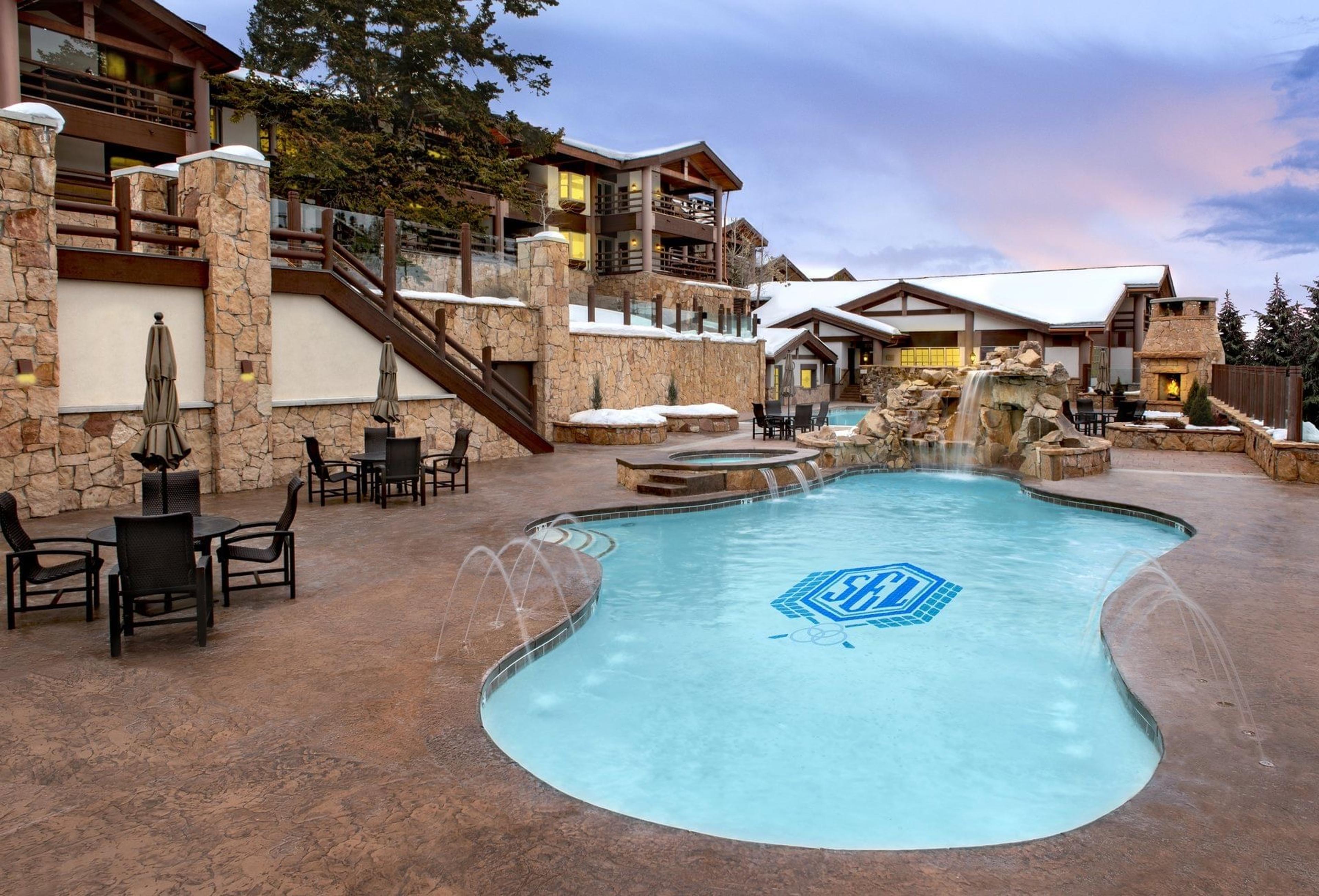 an outdoor pool with a rock waterfall feature at one end, loungers and umbrellas around it, lit by a pink evening sky