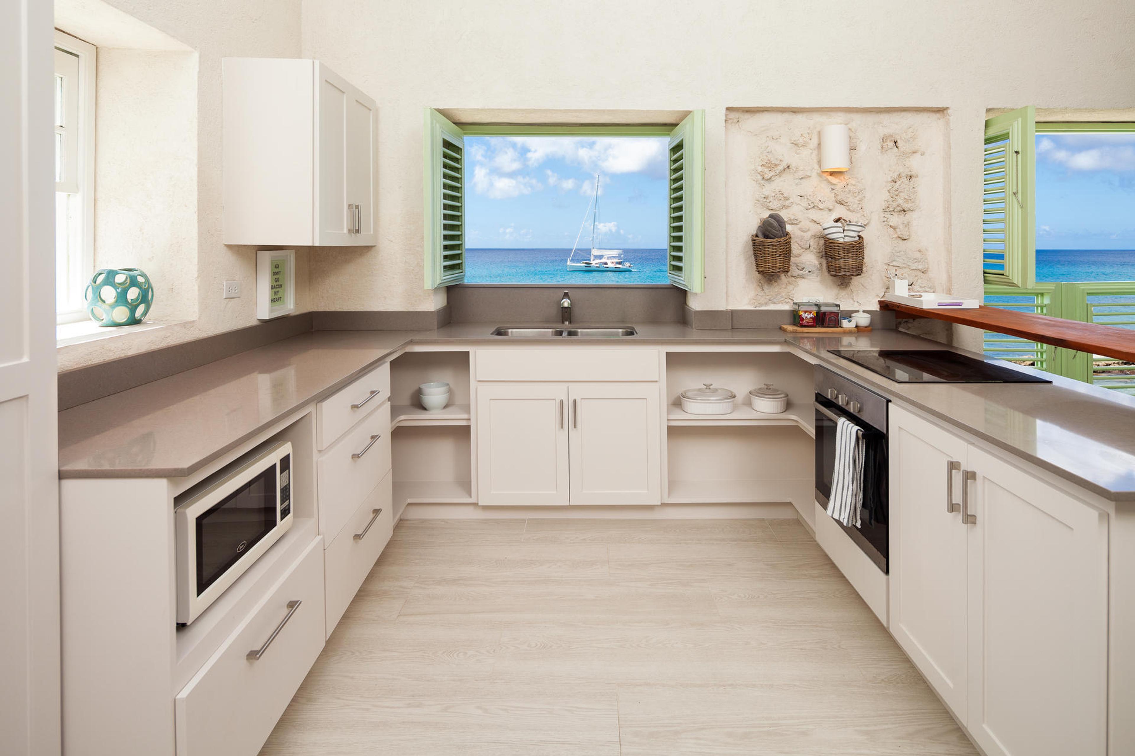A kitchen with white cabinets and a window open to the sea, a sailboat anchored offshore and green shutters either side.