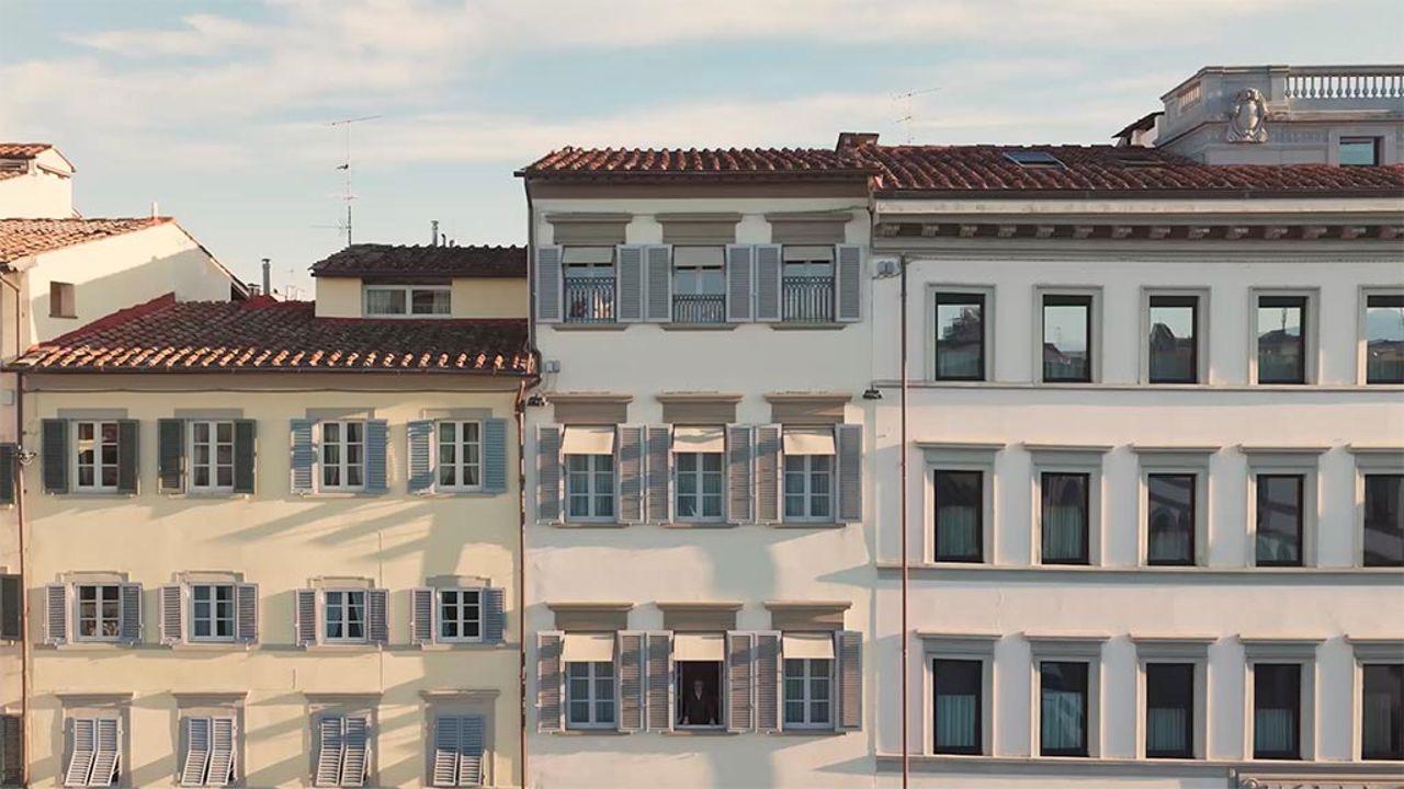 Three adjoining Florentine facades with grey shutters and terracotta roofs, seen from across the street in late afternoon light, with a person visible at a distance in one window.