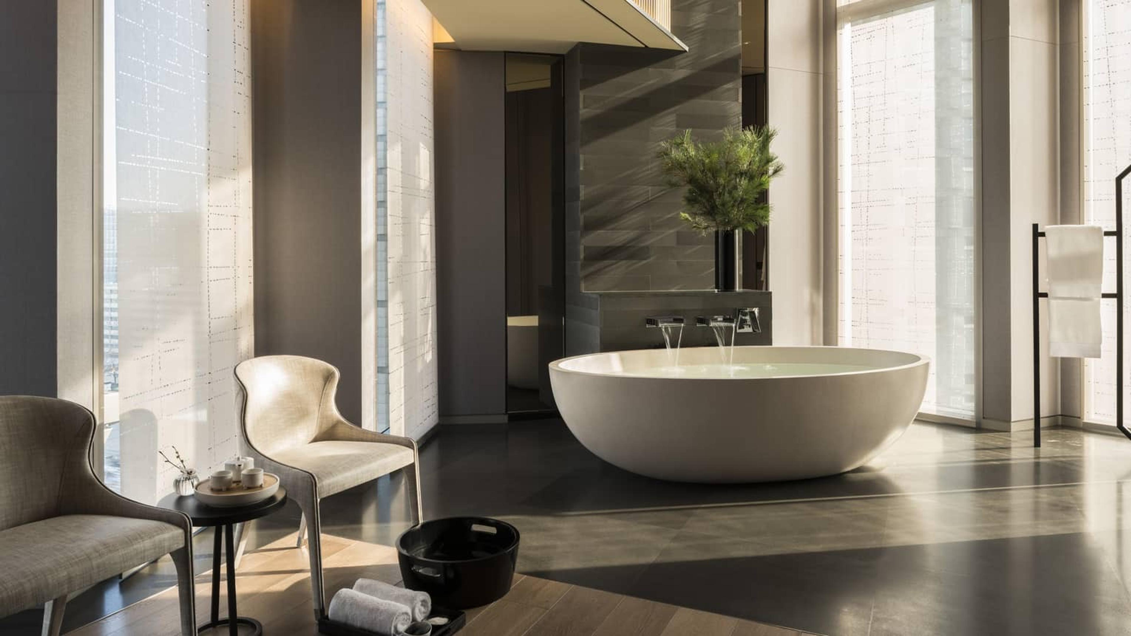 A round stone bathtub filling with water in a spa room, with two chairs, folded towels, and a small footbath nearby.