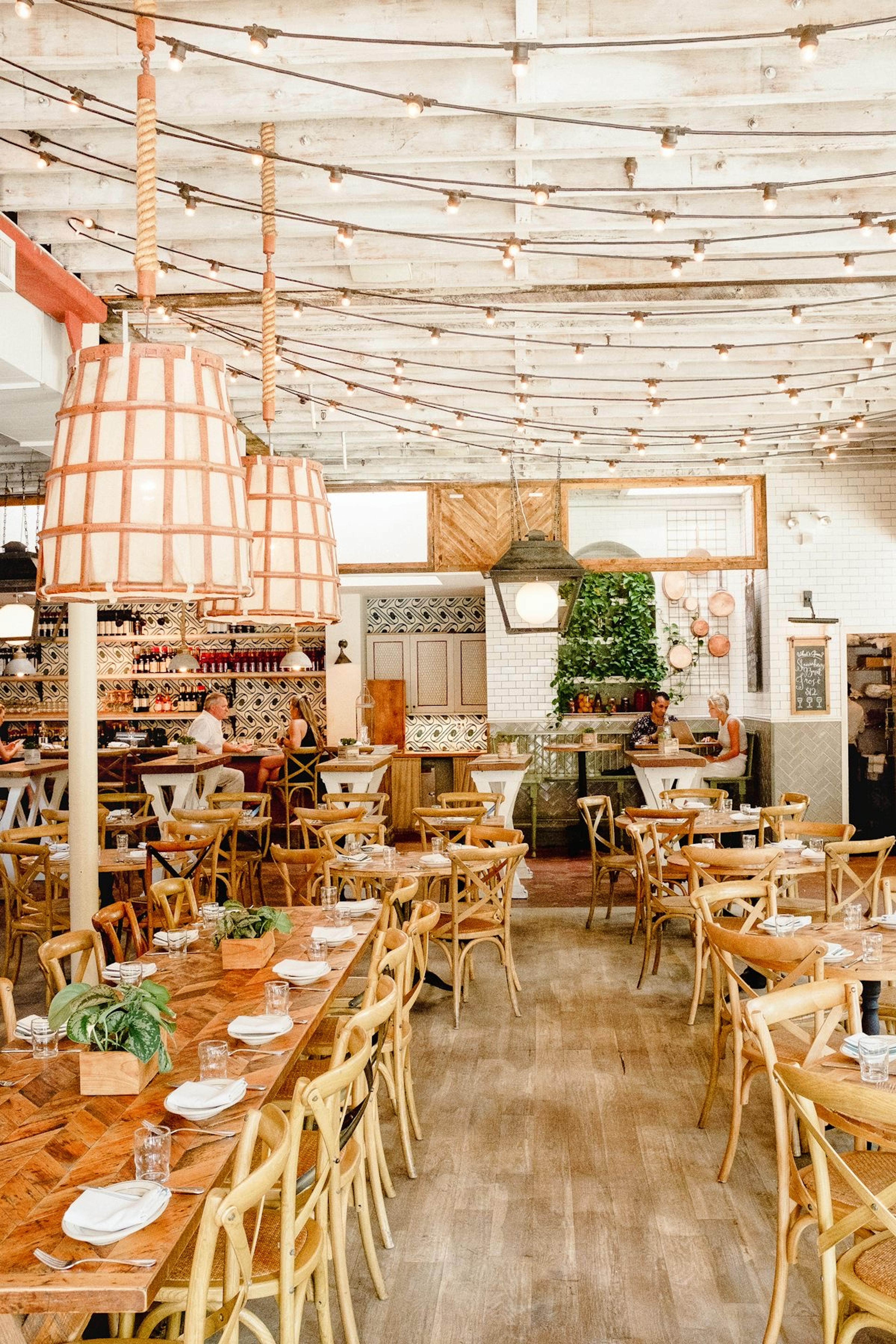 A bright corner dining room with wooden tables, hanging plants and woven pendant lamps