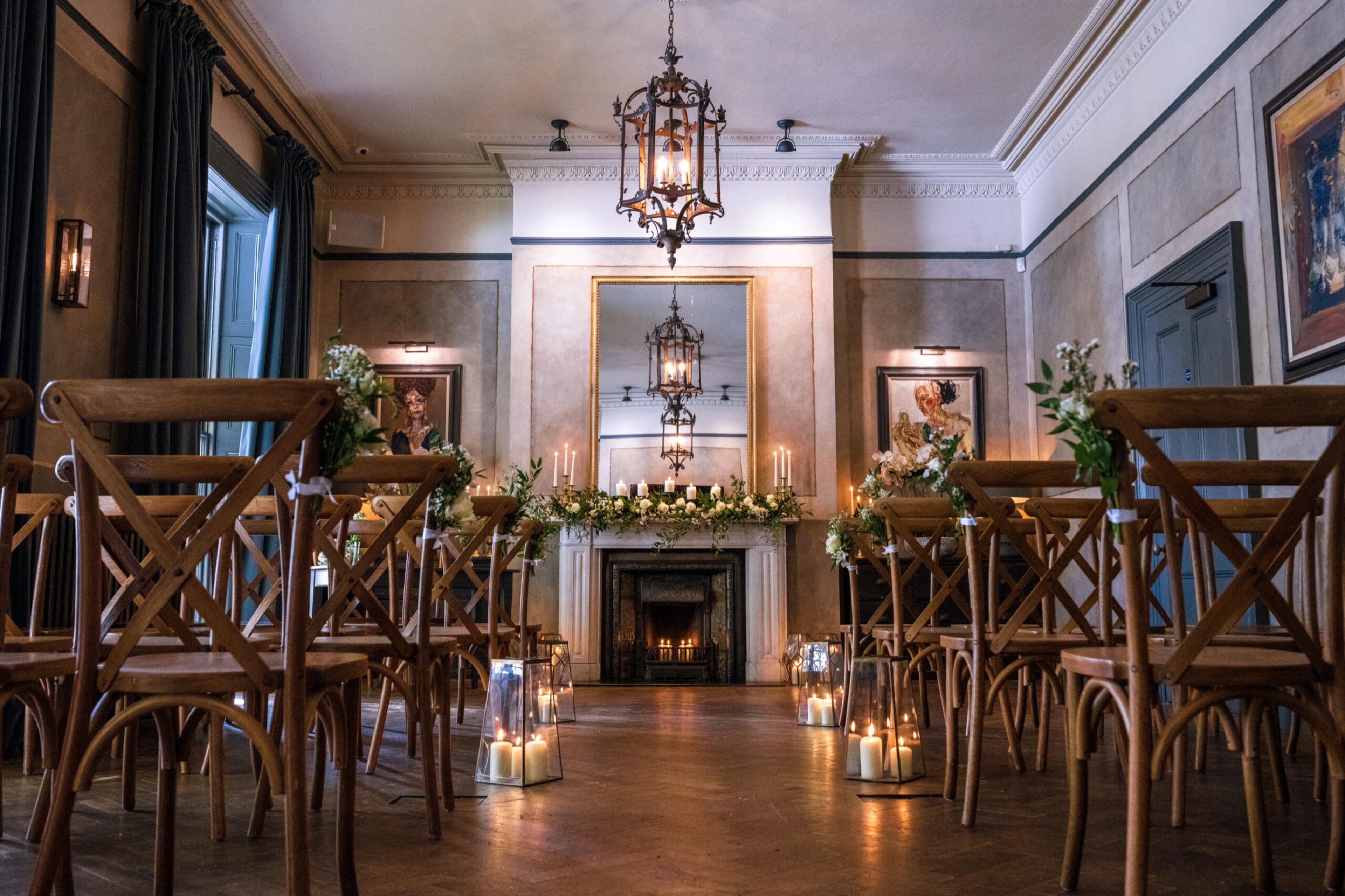 Rows of wooden chairs dressed with flowers face a lit fireplace and mirror in a wood-floored room, with lanterns of candles on the floor.