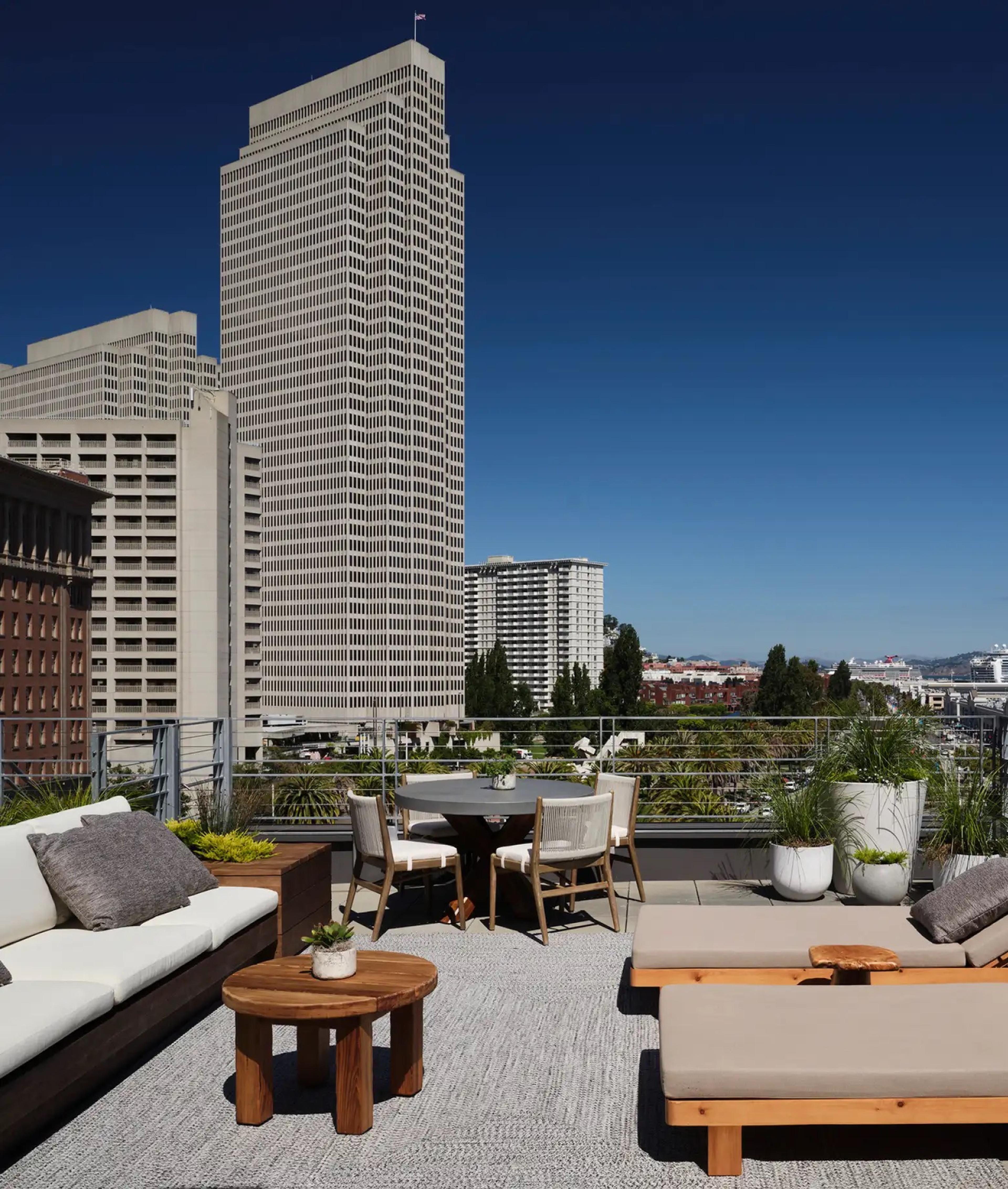 A rooftop terrace with a sofa, teak loungers and a round table, with downtown office towers against a clear blue sky.