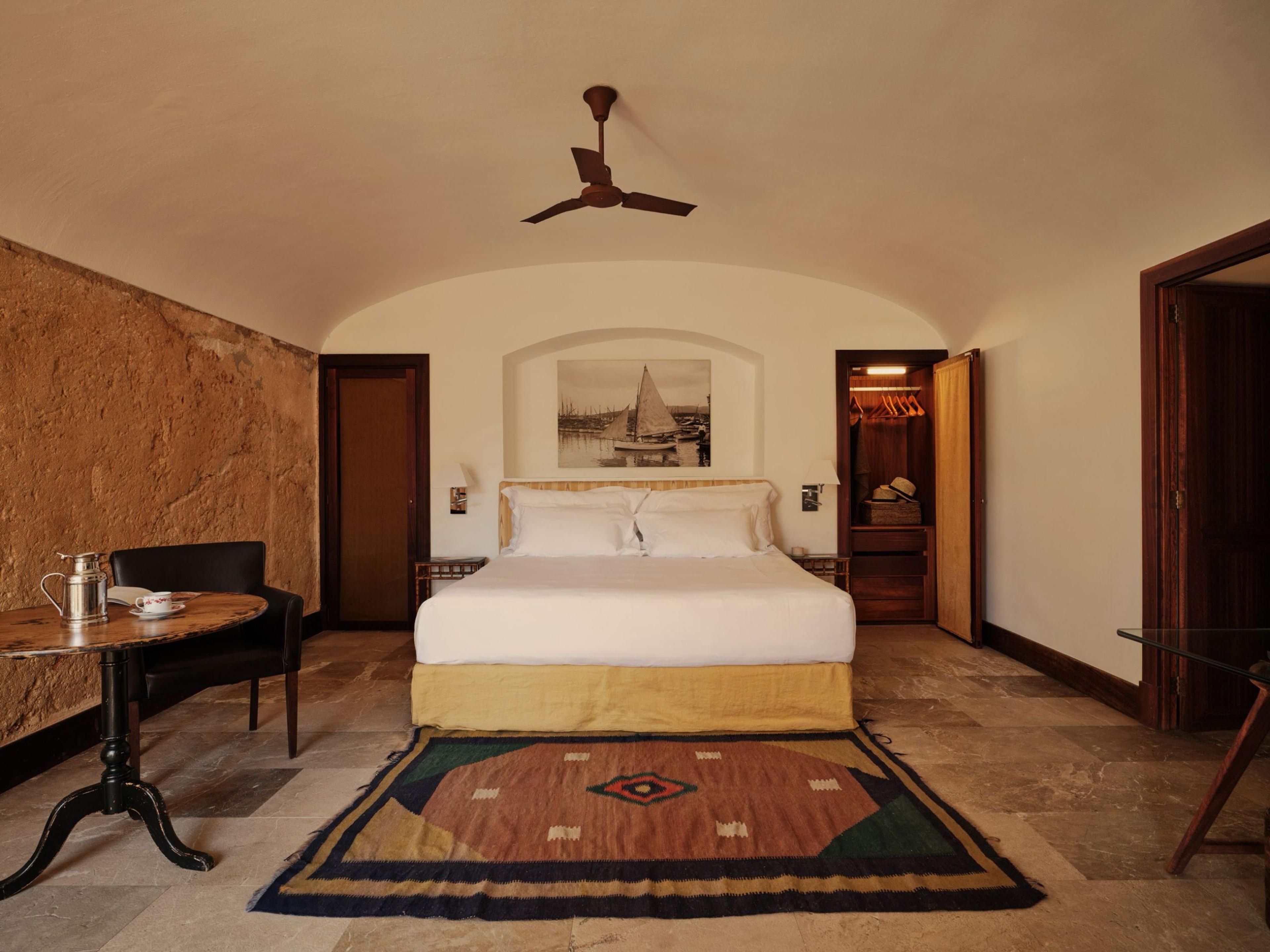 A bedroom with a vaulted stone ceiling, a ceiling fan, a white bed with a mustard-colored base, and a patterned rug on a tiled floor.