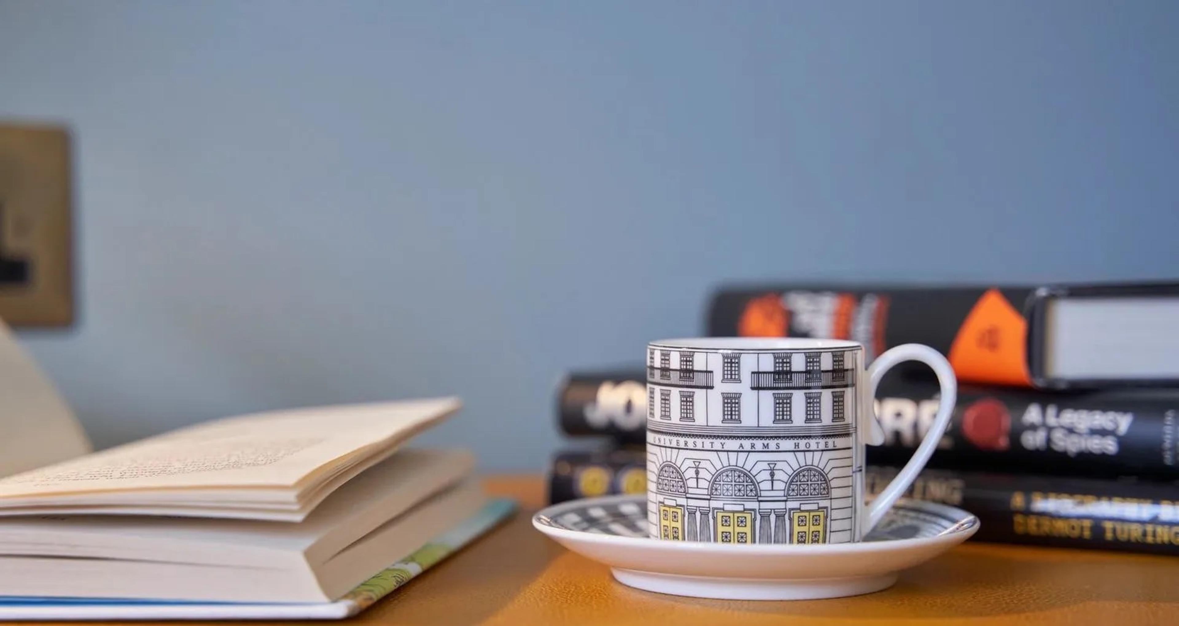 a teacup and saucer printed with an illustration of the hotel's facade, beside an open book and a stack of books on a wooden table against a pale blue wall