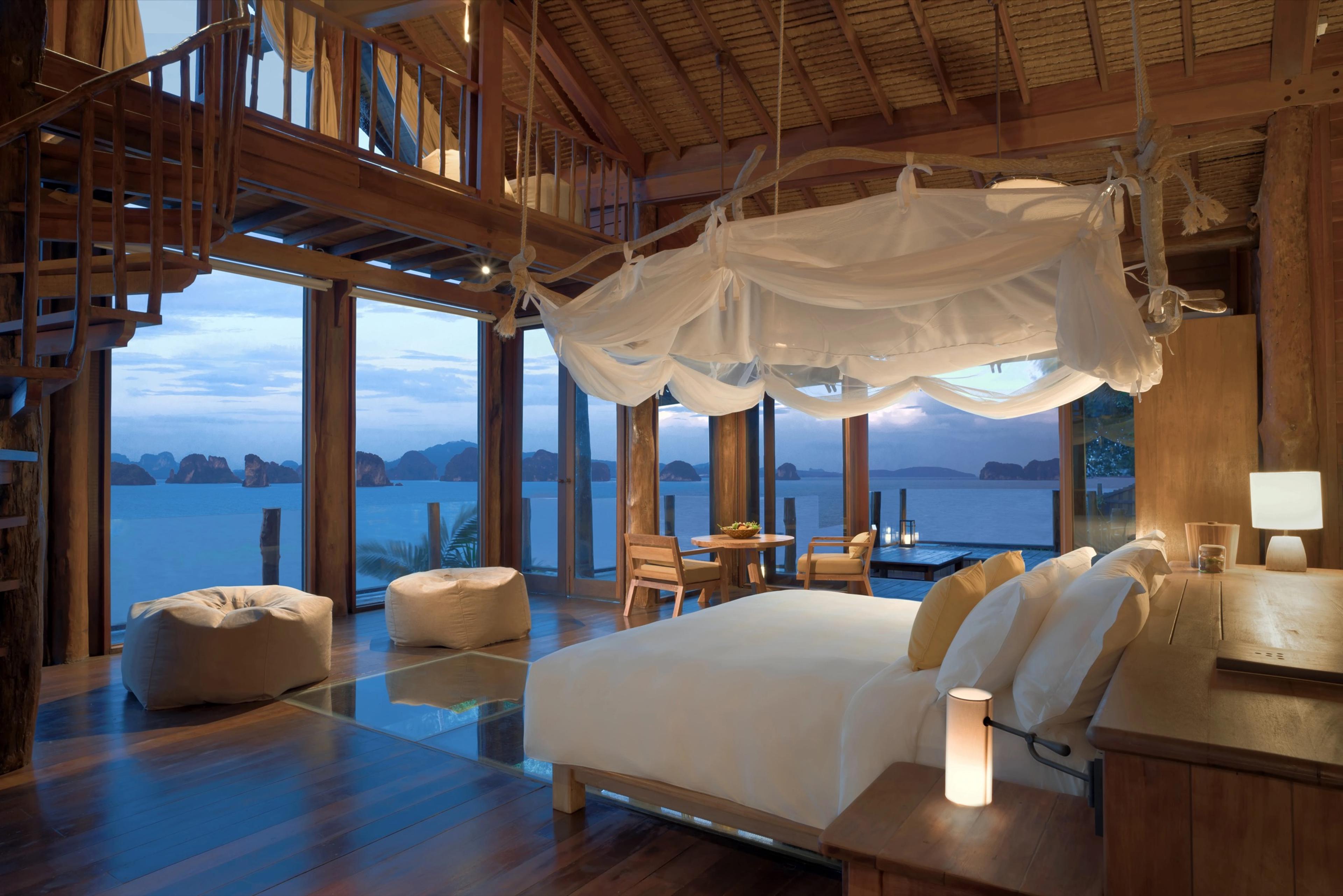 A wood-framed bedroom with a draped canopy bed, a spiral staircase to a loft, and floor-to-ceiling windows looking out over the sea at dusk.