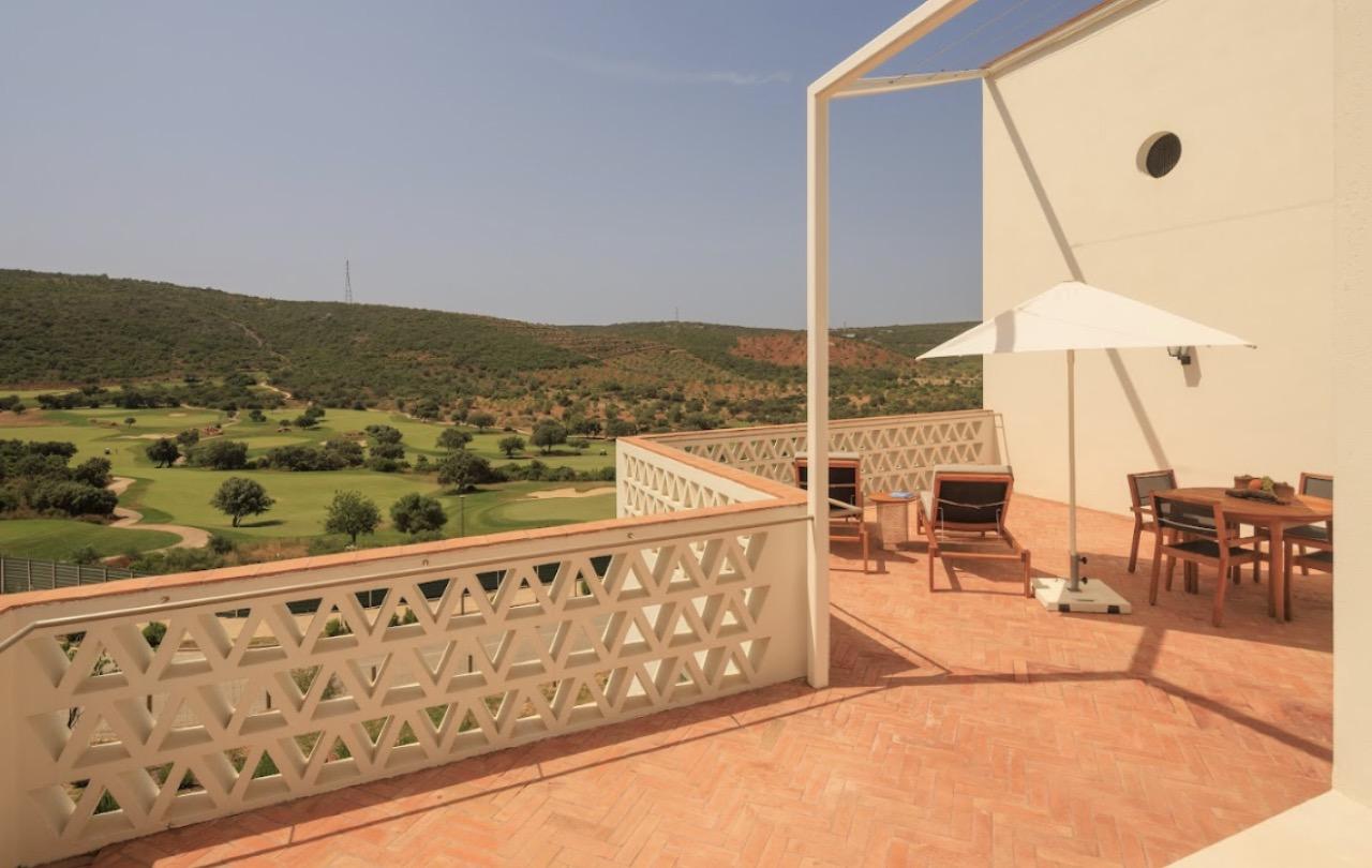 A tiled terrace with a dining table, chairs and a market umbrella against a whitewashed wall, overlooking a golf course valley.