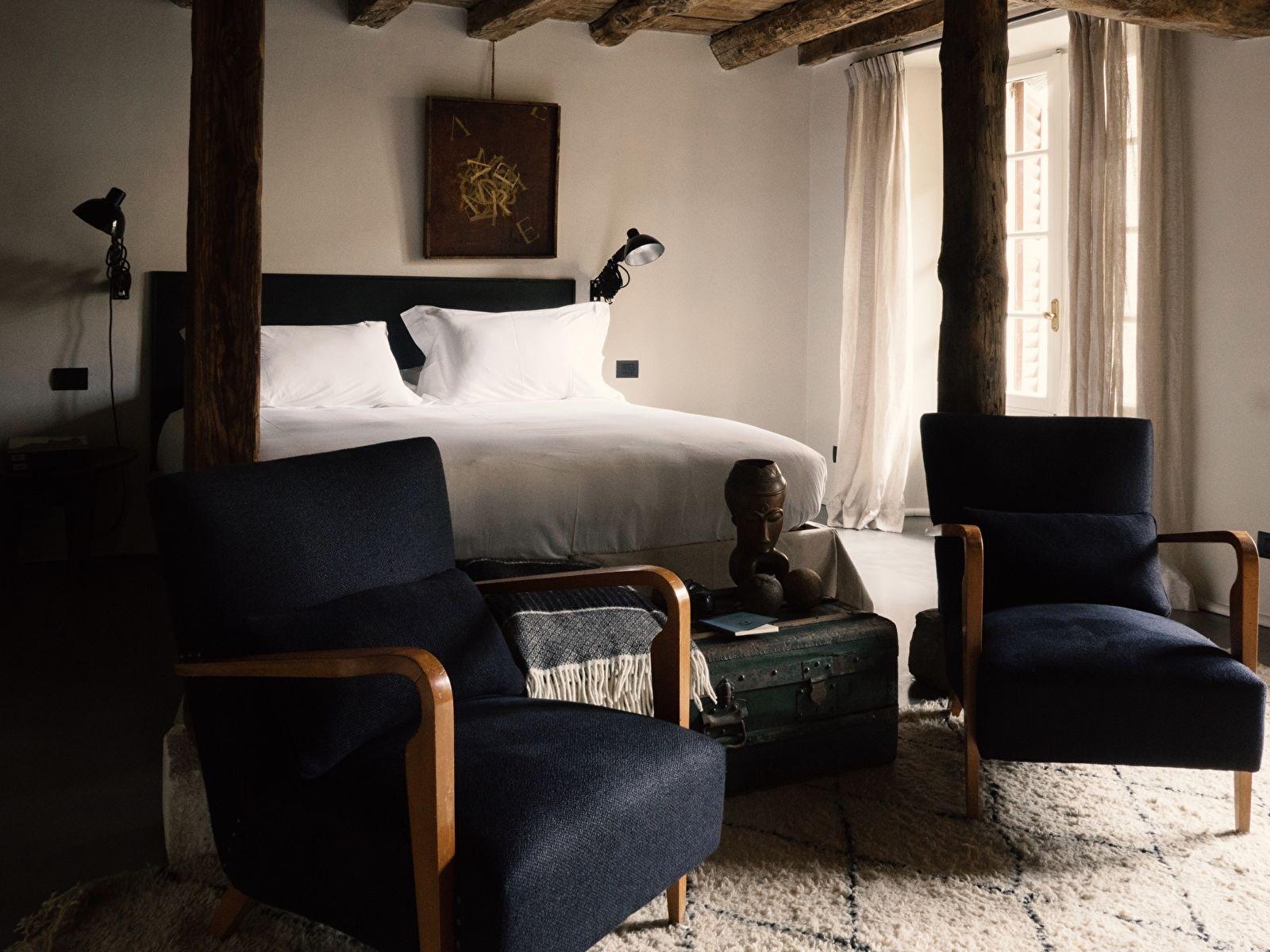 a bed between wooden ceiling posts, flanked by two navy armchairs, with a leather trunk and a carved wooden figure in front