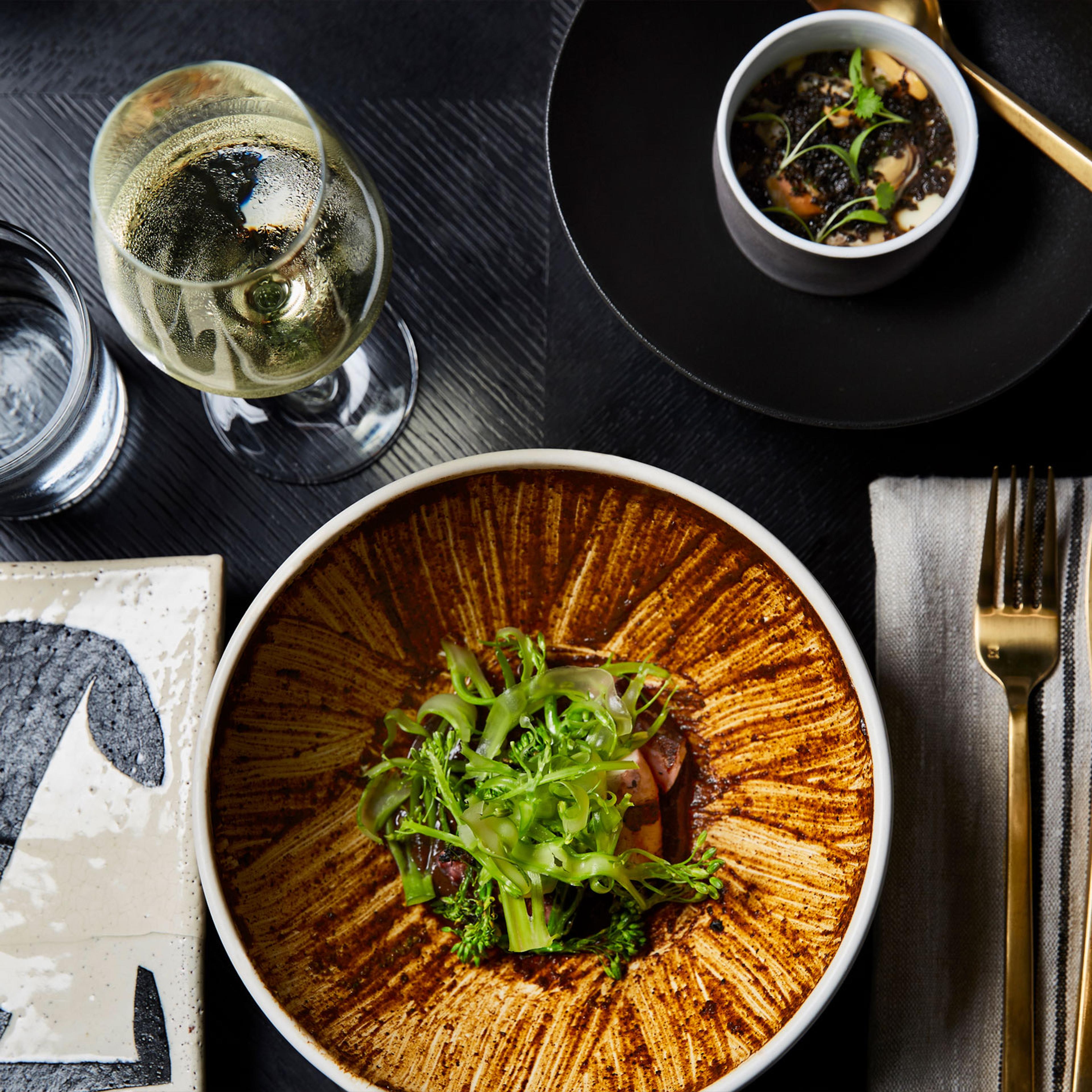 An overhead view of a plated dish with pickled vegetables and herbs beside a small bowl of black grains, a glass of white wine and cutlery on a dark table.