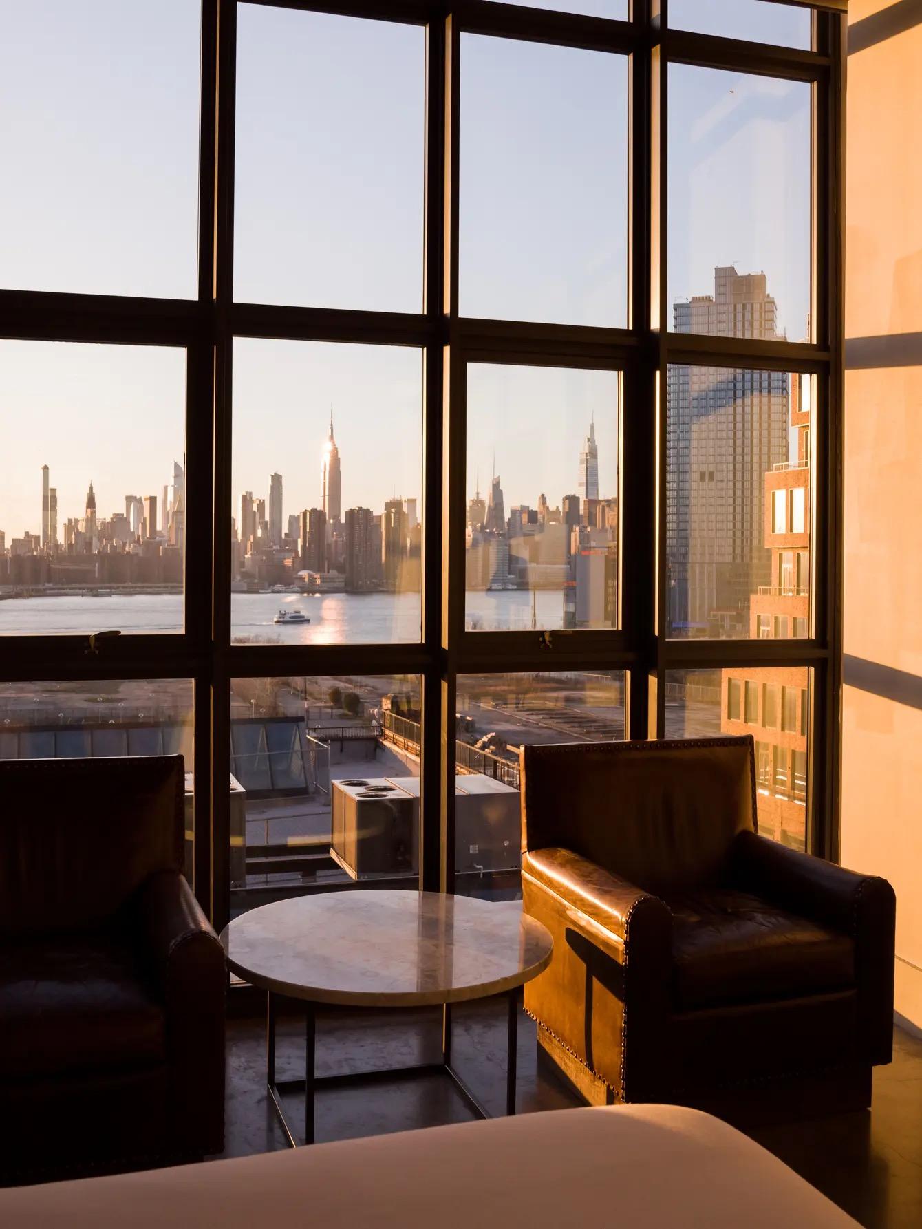 Two leather armchairs around a round marble table in front of a floor to ceiling window, golden evening light on the Manhattan skyline across the water.