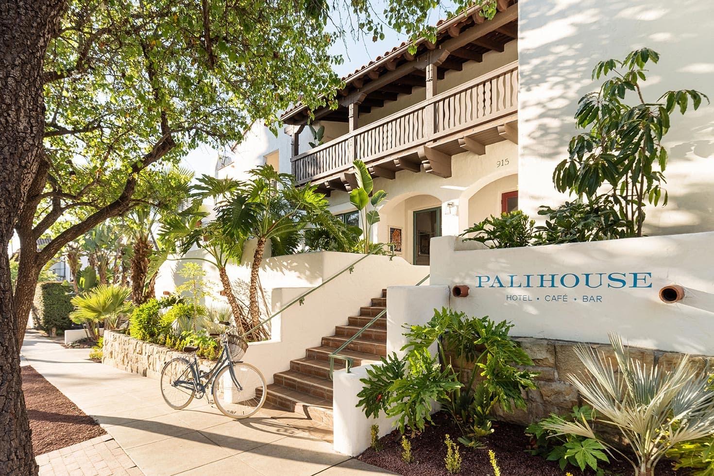Stone steps lead up from a tree-lined sidewalk to a tiled entrance beneath a wooden balcony, with a bicycle parked against the low wall and the words Palihouse Hotel Cafe Bar painted on a sign.