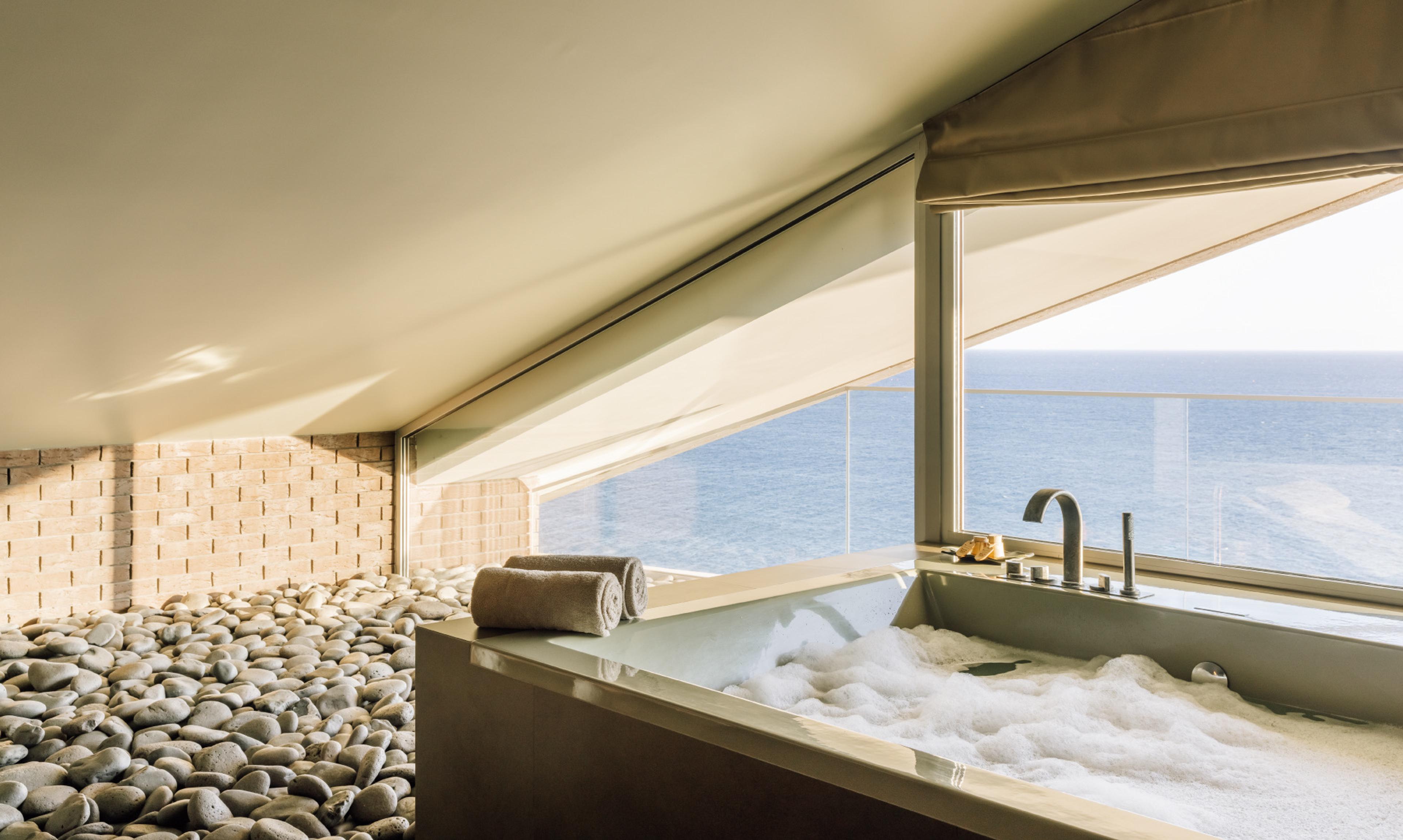 A built in bathtub full of bubbles beside a pebbled floor and a window onto the sea.