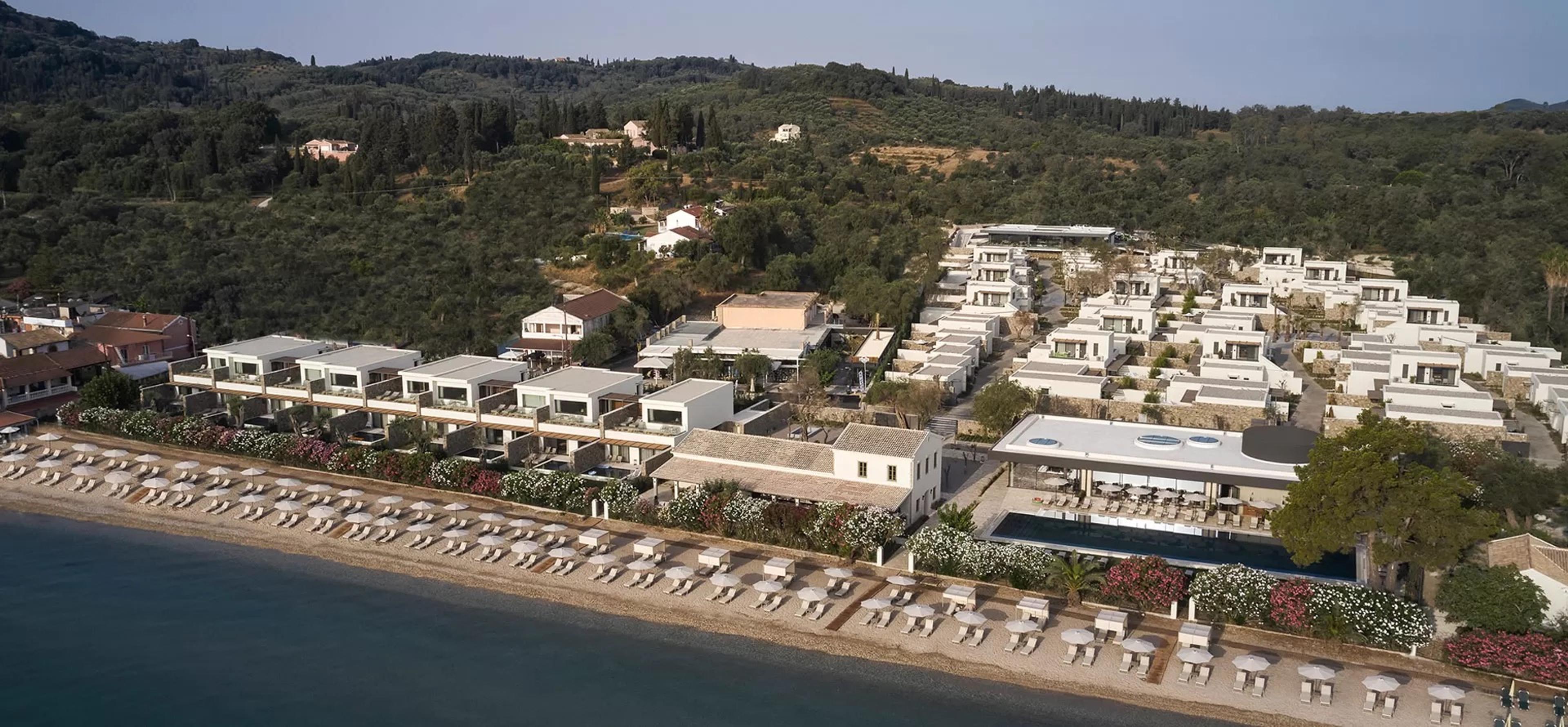 An aerial view of a hotel complex of white buildings and pools set among green hillside, with a beach and the sea below.