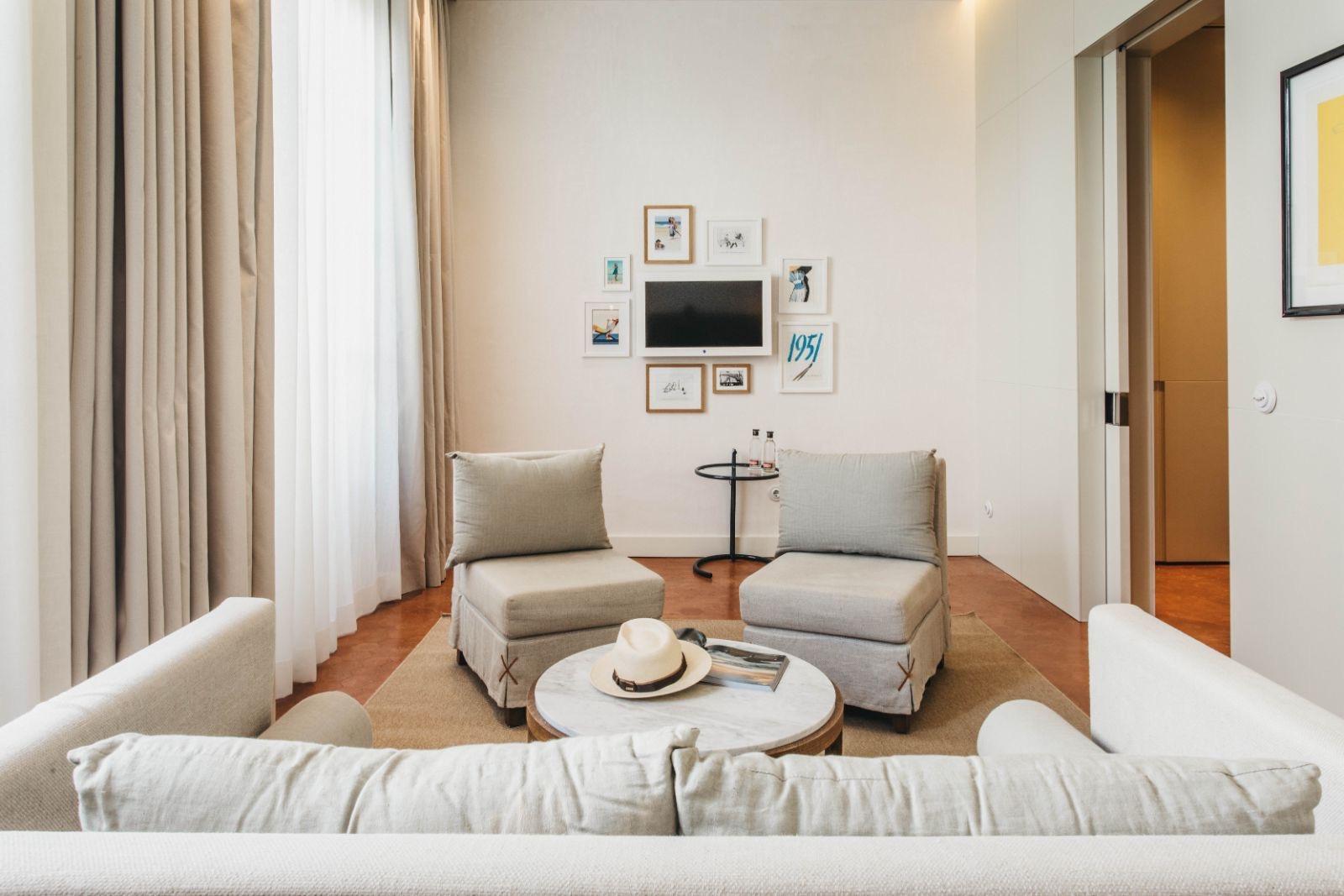 A sitting area with two armchairs and a round coffee table in front of a sofa, framed pictures and a television on the wall, and a straw hat on the table.