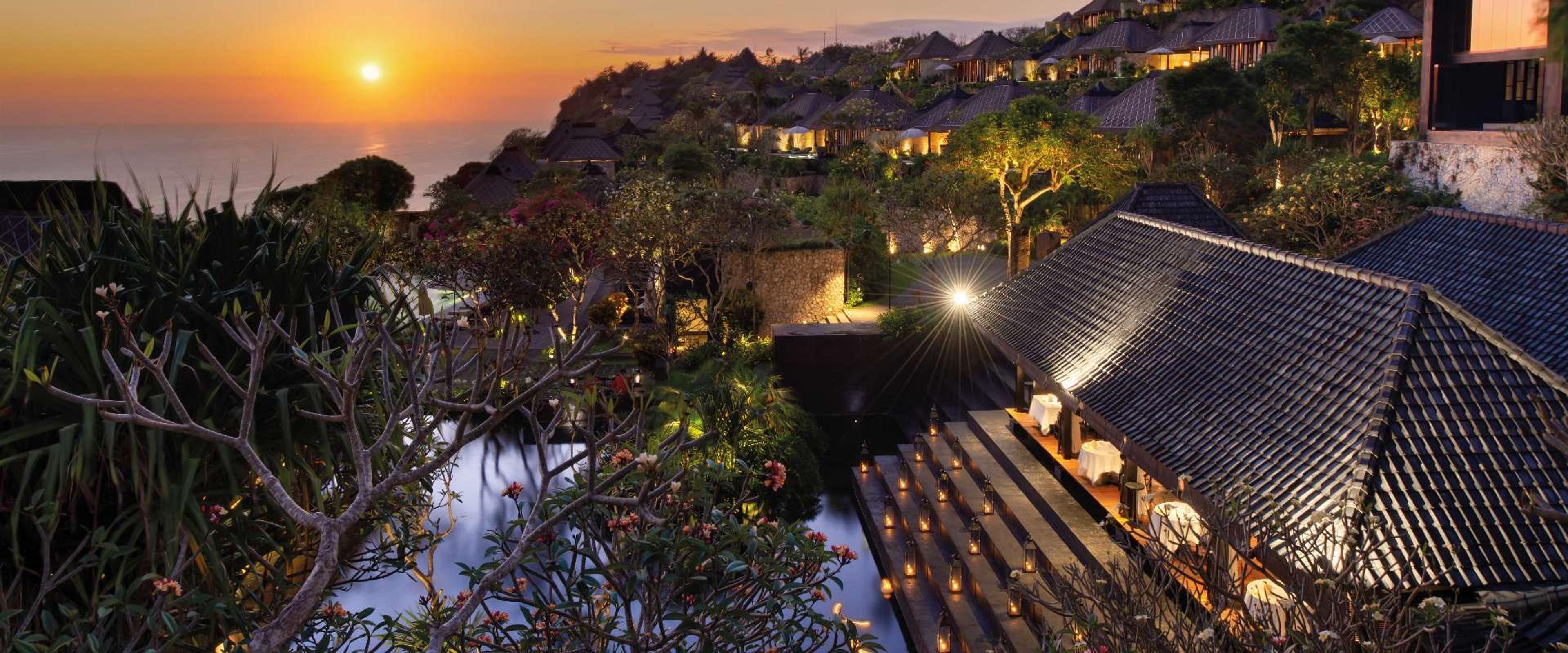 The resort at sunset, lit villas stepping down the cliff and a lantern-lined stair to the restaurant