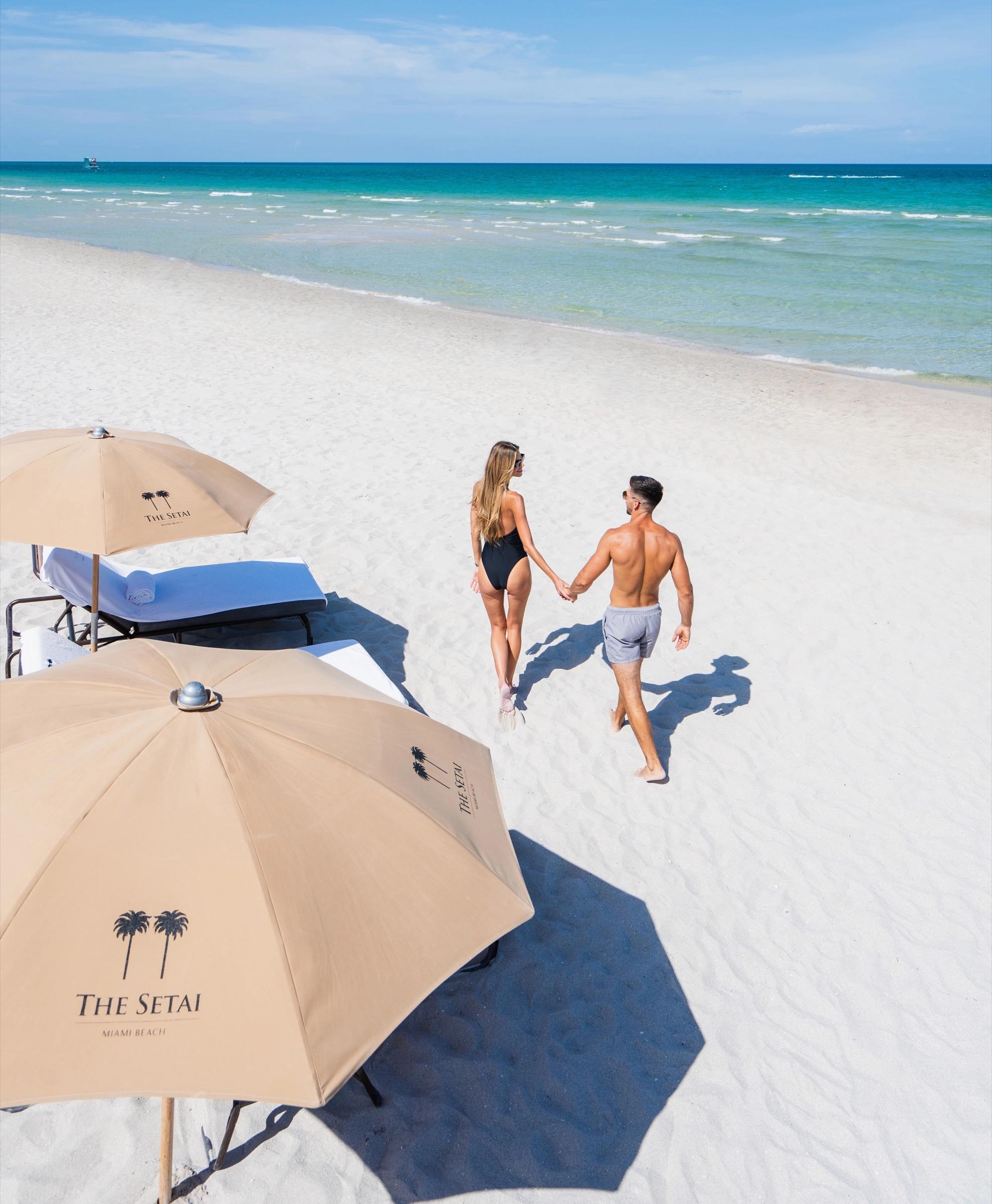 A couple hold hands and walk away from the camera along an empty white sand beach, beach umbrellas in the foreground.