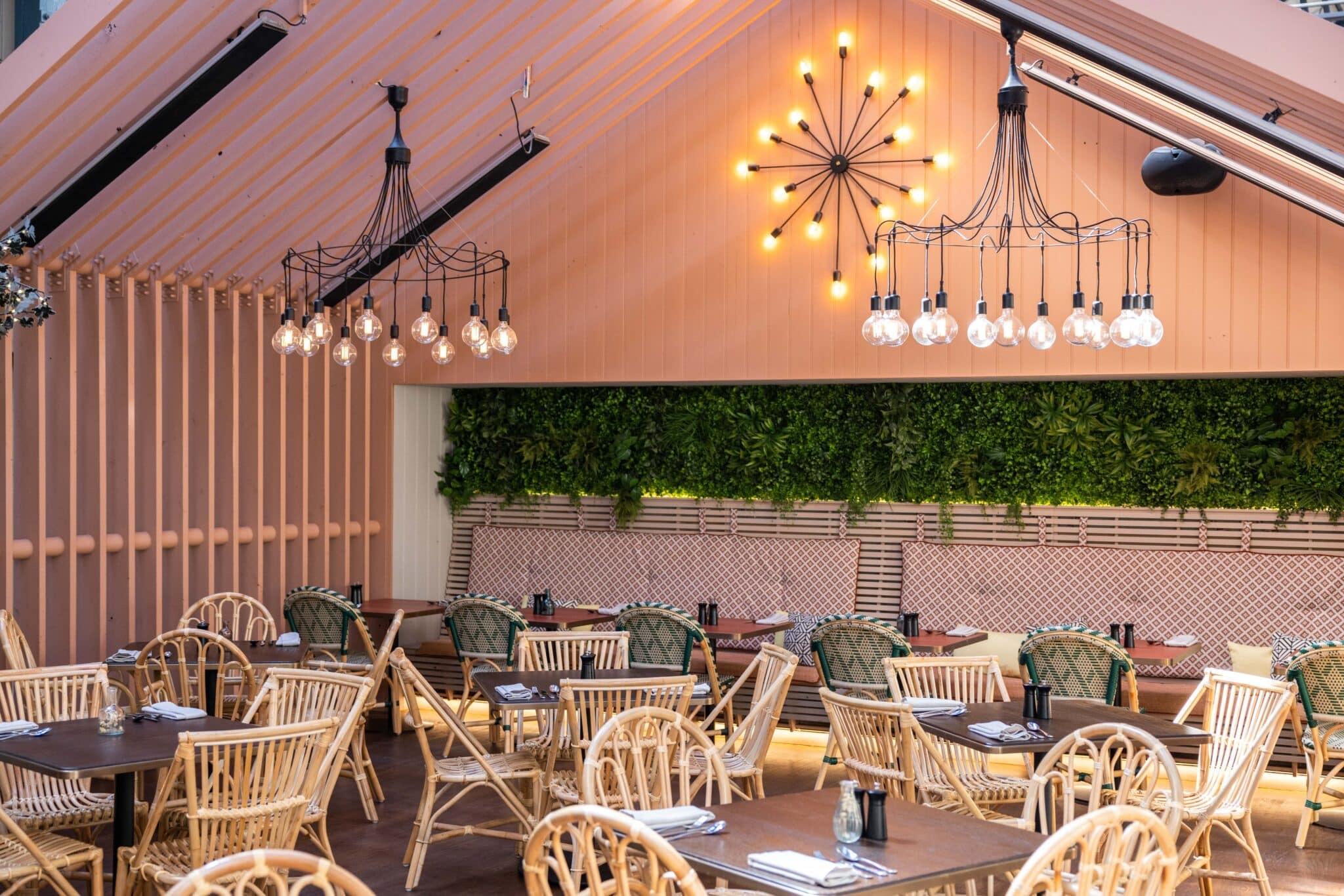 An open-sided terrace dining room with a pink pitched roof, wrought-iron chandeliers, a green wall and rattan chairs at set tables.