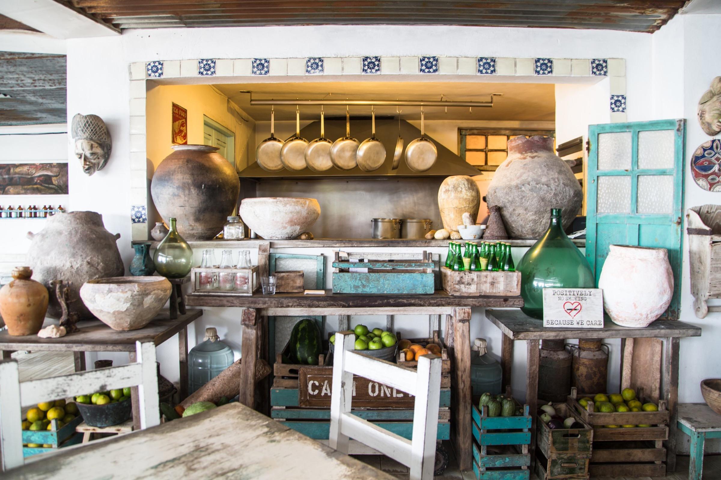 A whitewashed room of shelves stacked with clay pots and copper pans, crates of fruit below