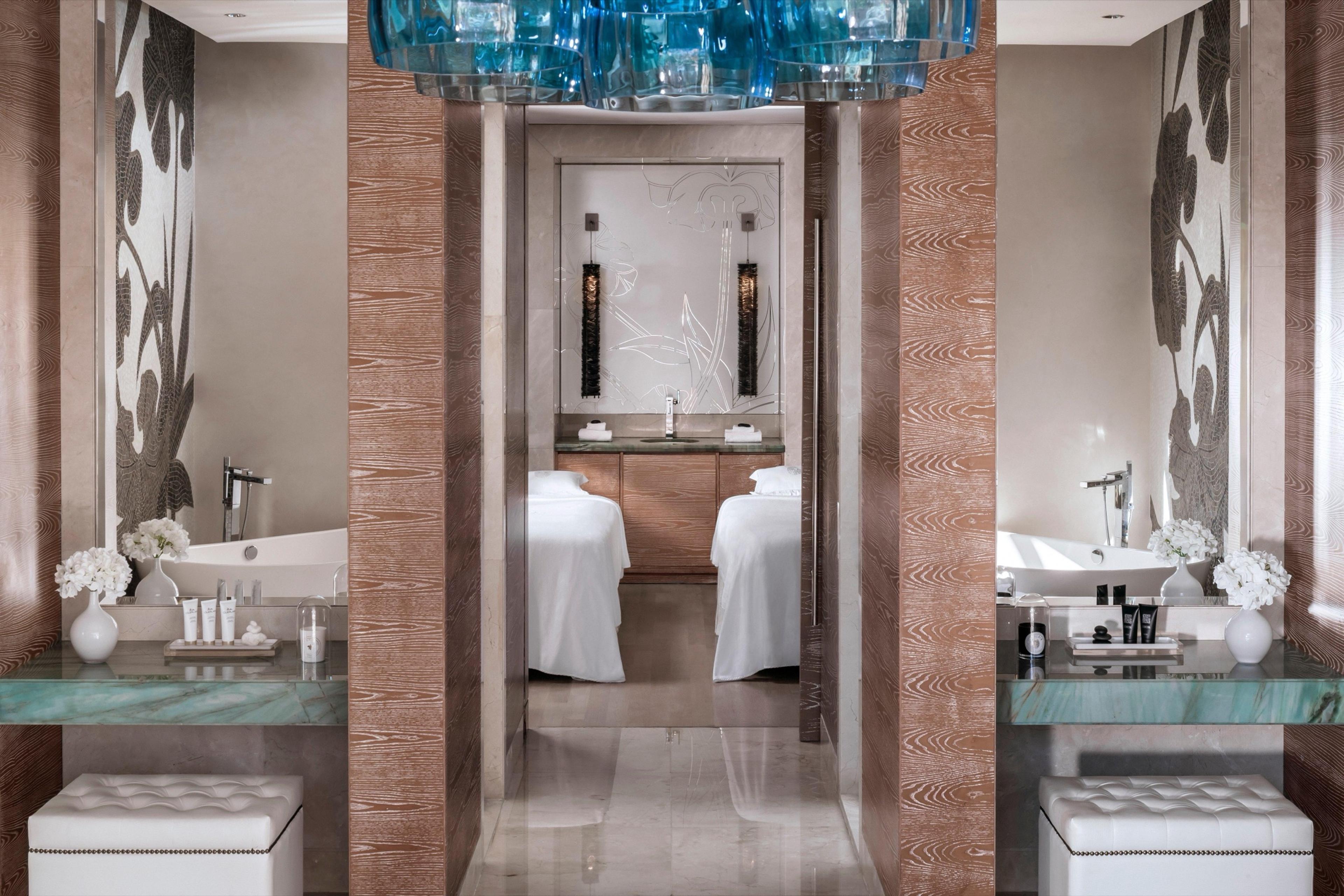 A spa suite with two treatment beds is reflected in mirrors on either side, framed by a blue glass chandelier and wood-paneled columns.