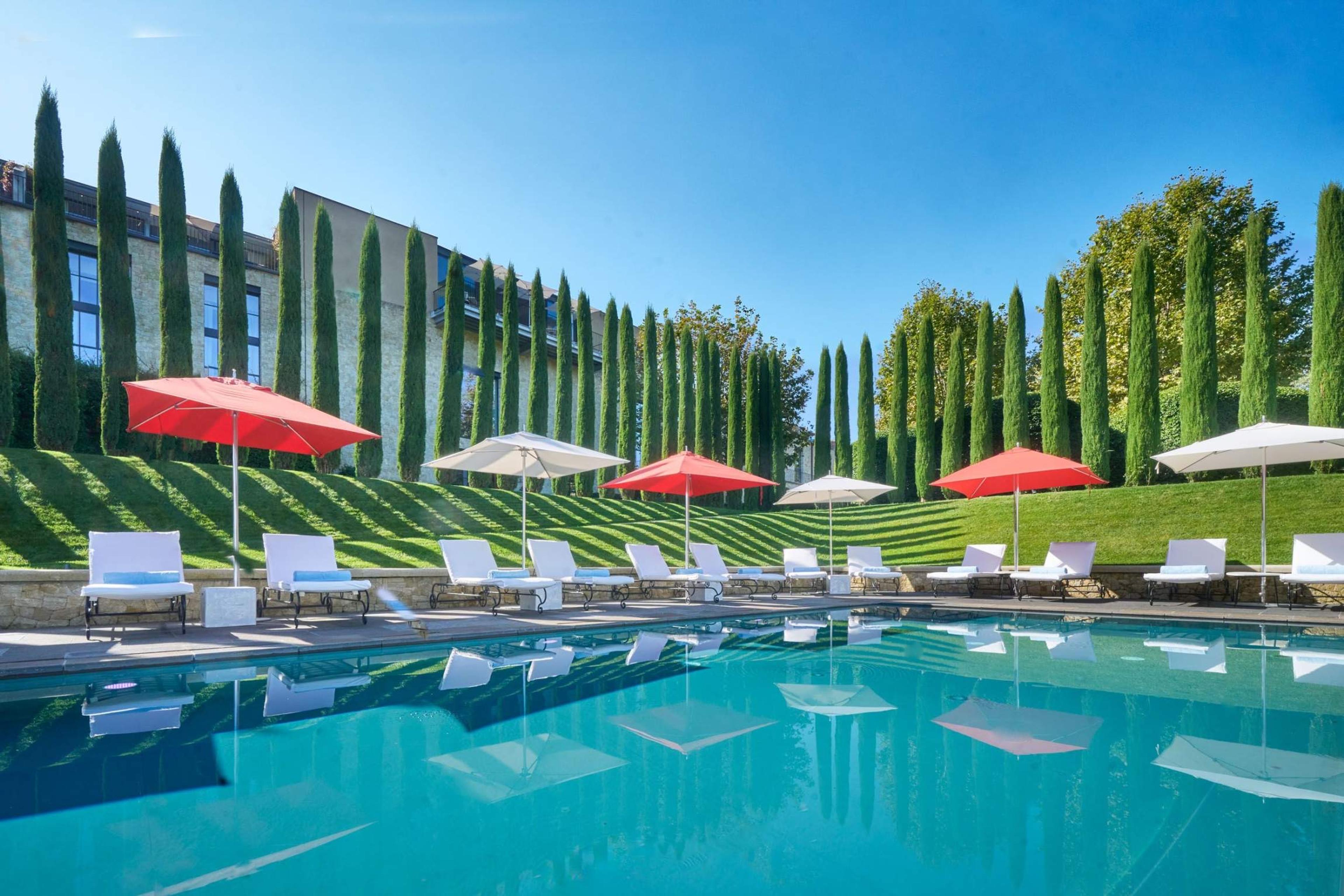 A rectangular pool beside a long glass-fronted building, with red and white sun umbrellas on white loungers and a row of cypress trees on the slope behind.