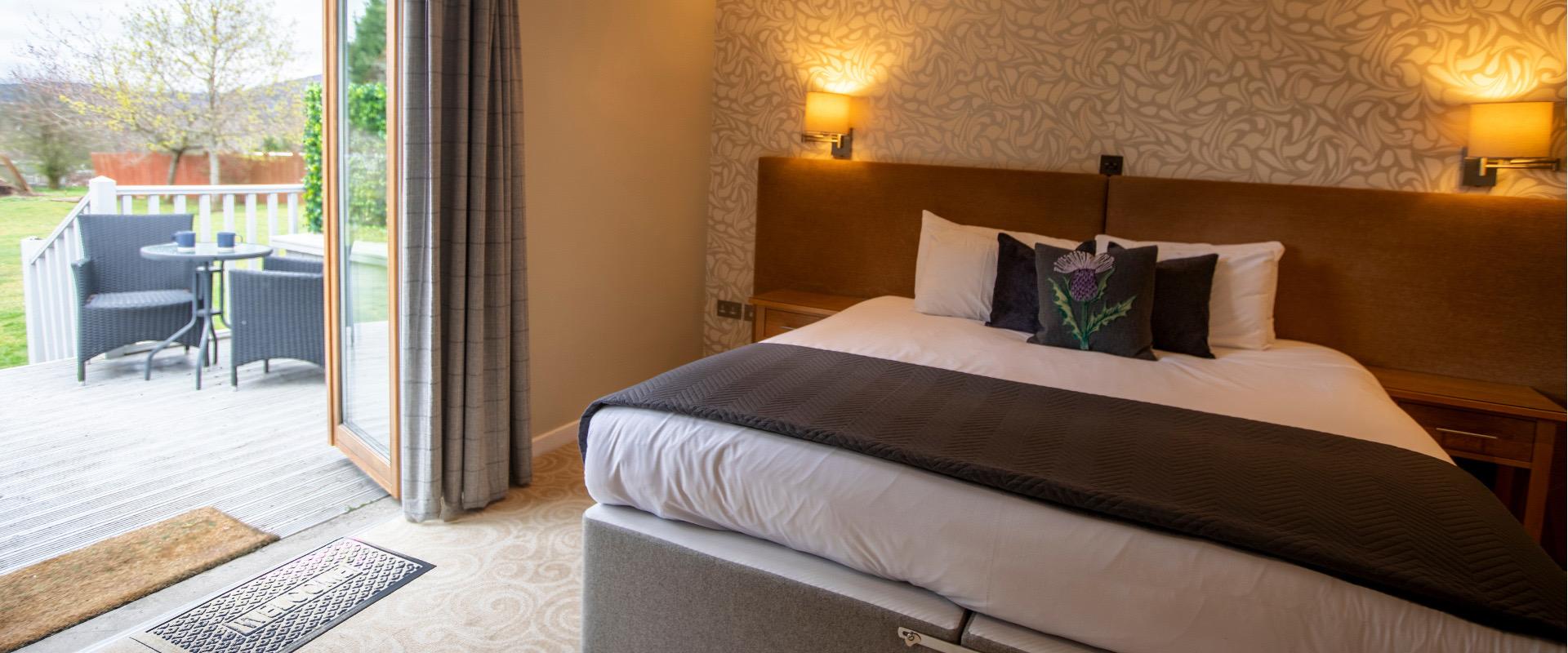 A hotel bedroom with a bed dressed in grey and white linen and a thistle embroidered cushion, patio doors open onto a deck with two chairs and a garden beyond.