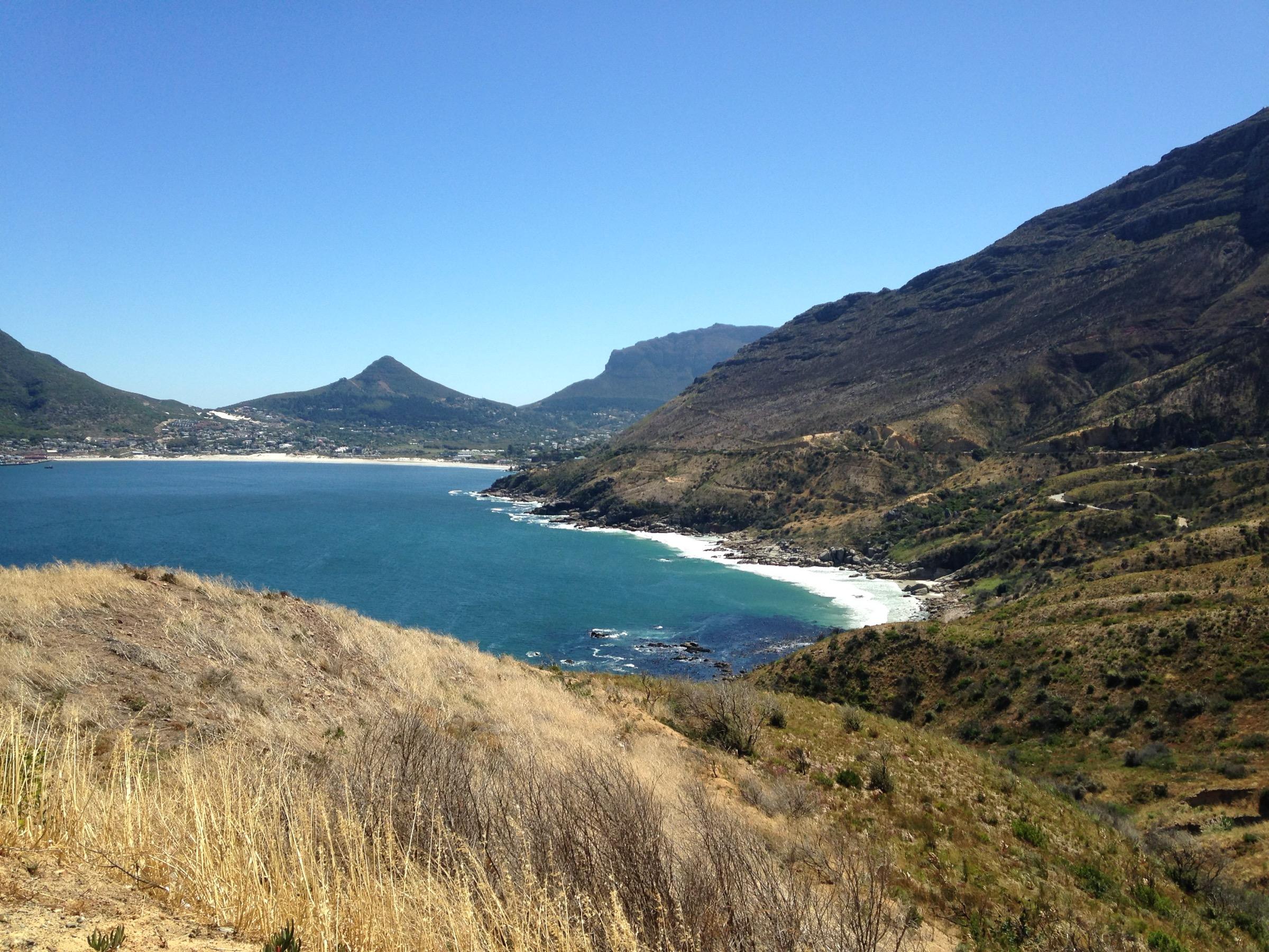 A blue bay with a white beach under mountains, seen from a dry, grassy hillside