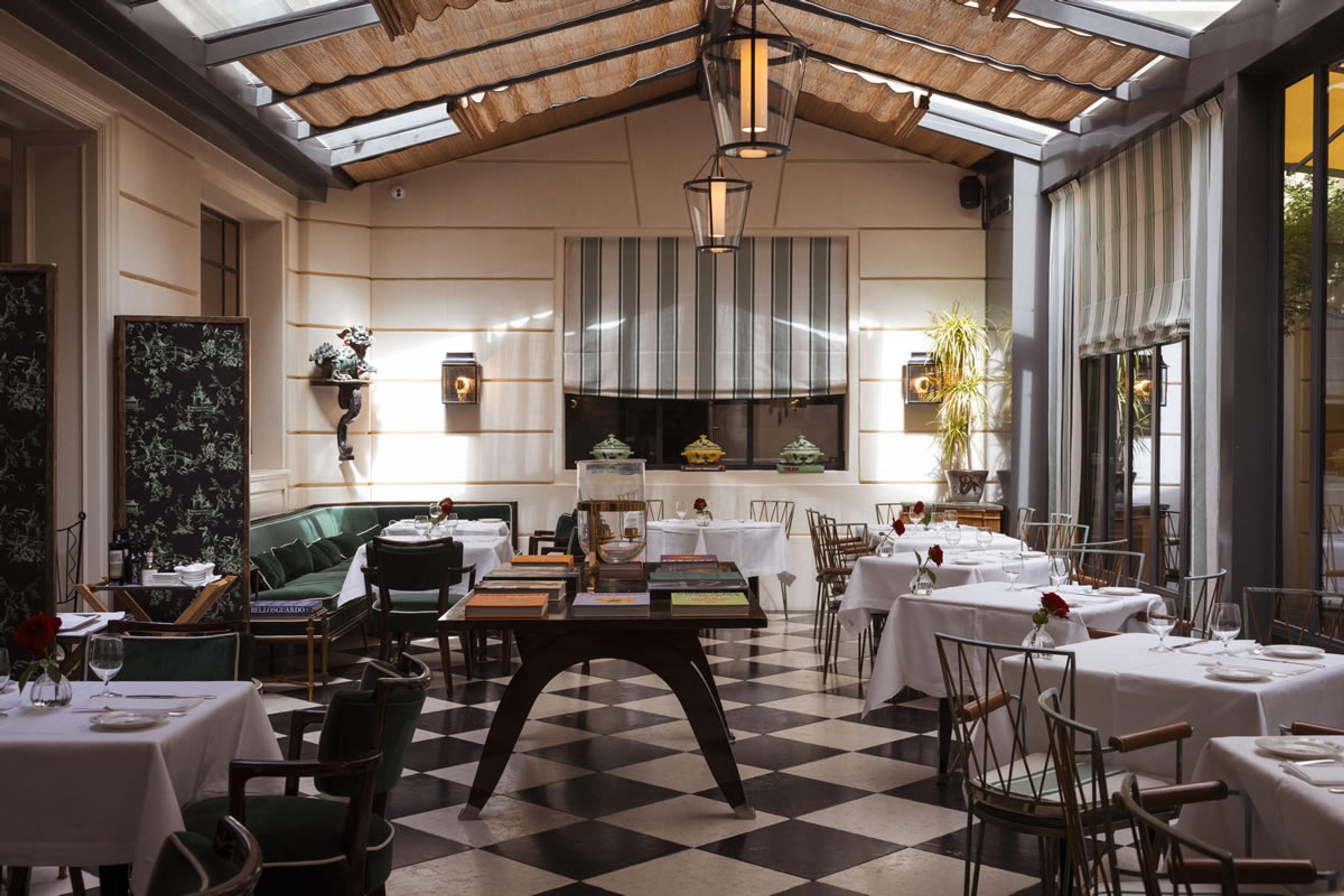 A glass-roofed dining room with white-clothed tables, wicker and metal chairs, a hanging lantern, and striped blinds at the windows.