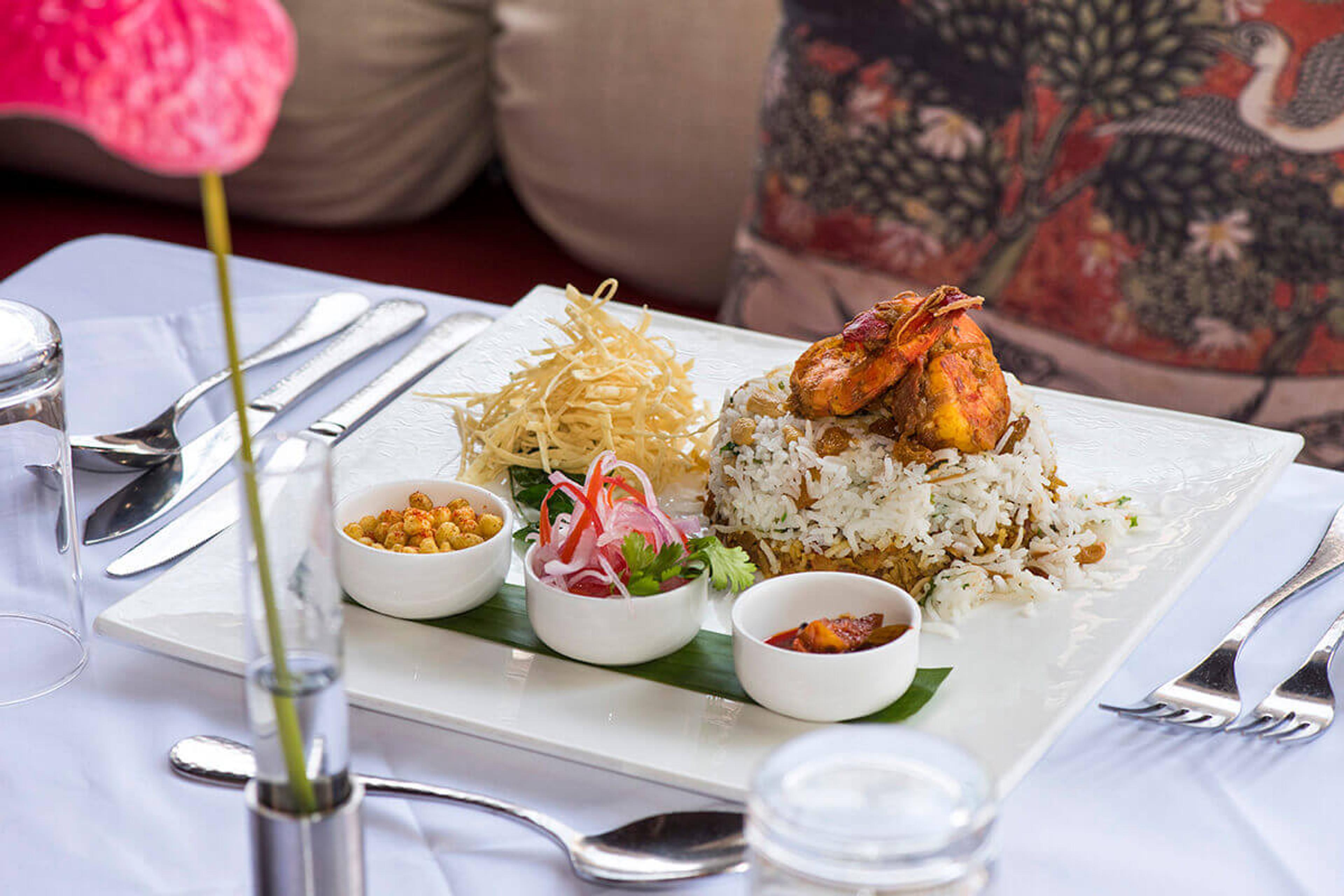 A plated dish of rice topped with grilled prawns, served with small bowls of chutney, pickle and salad on a white tablecloth.