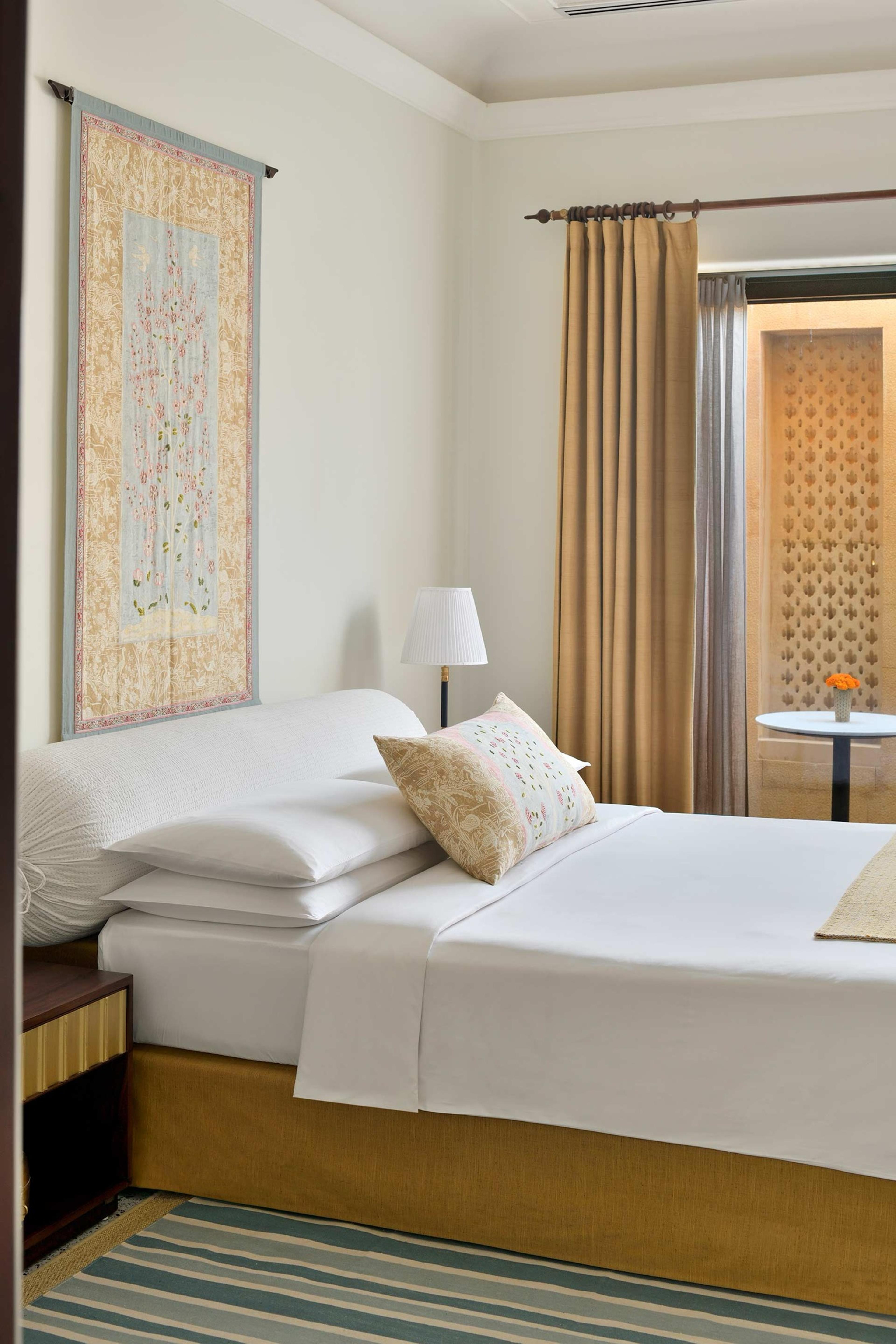 a made-up bed with white linen and bolster pillows, a tapestry on the wall behind and a striped rug on the floor
